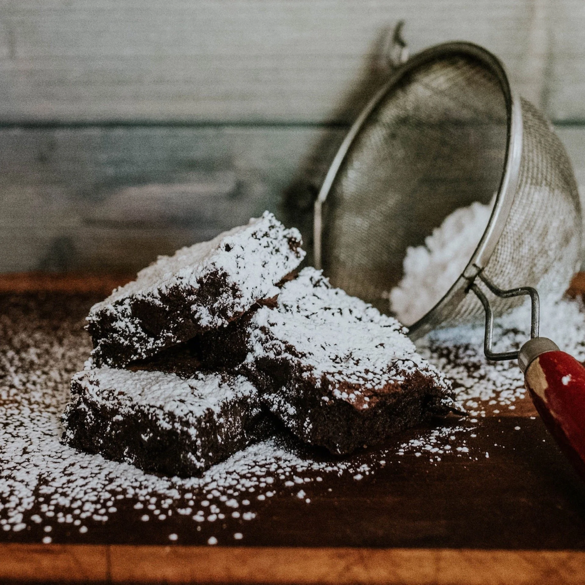 A close-up of a baking tray filled with chocolate chip cookies dusted with powdered sugar, resting on a wooden surface with a metal sieve nearby.