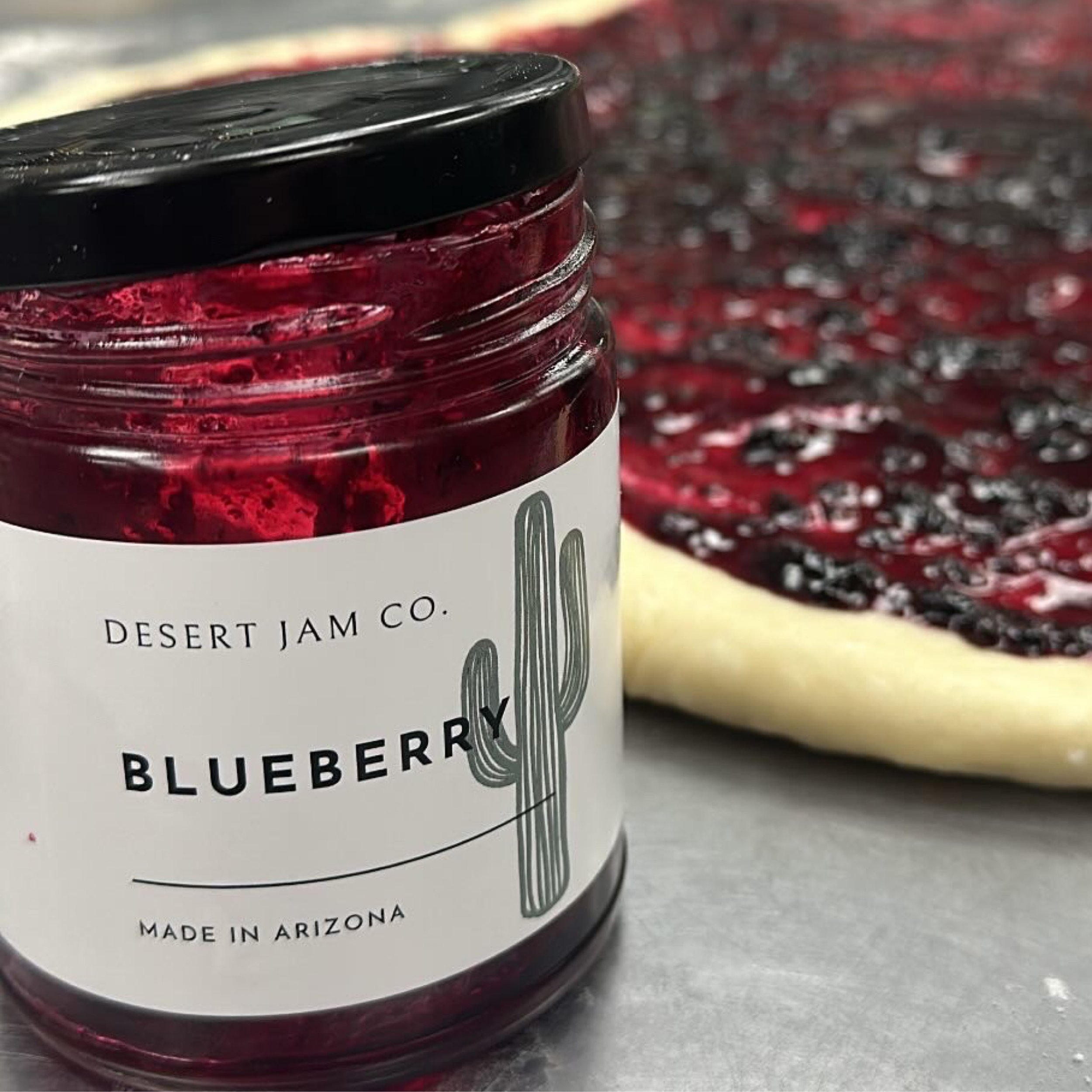 Close-up of a jar of berry jam with a black lid, placed on a gray surface, with a blurred background of the same jam spread on a plate.