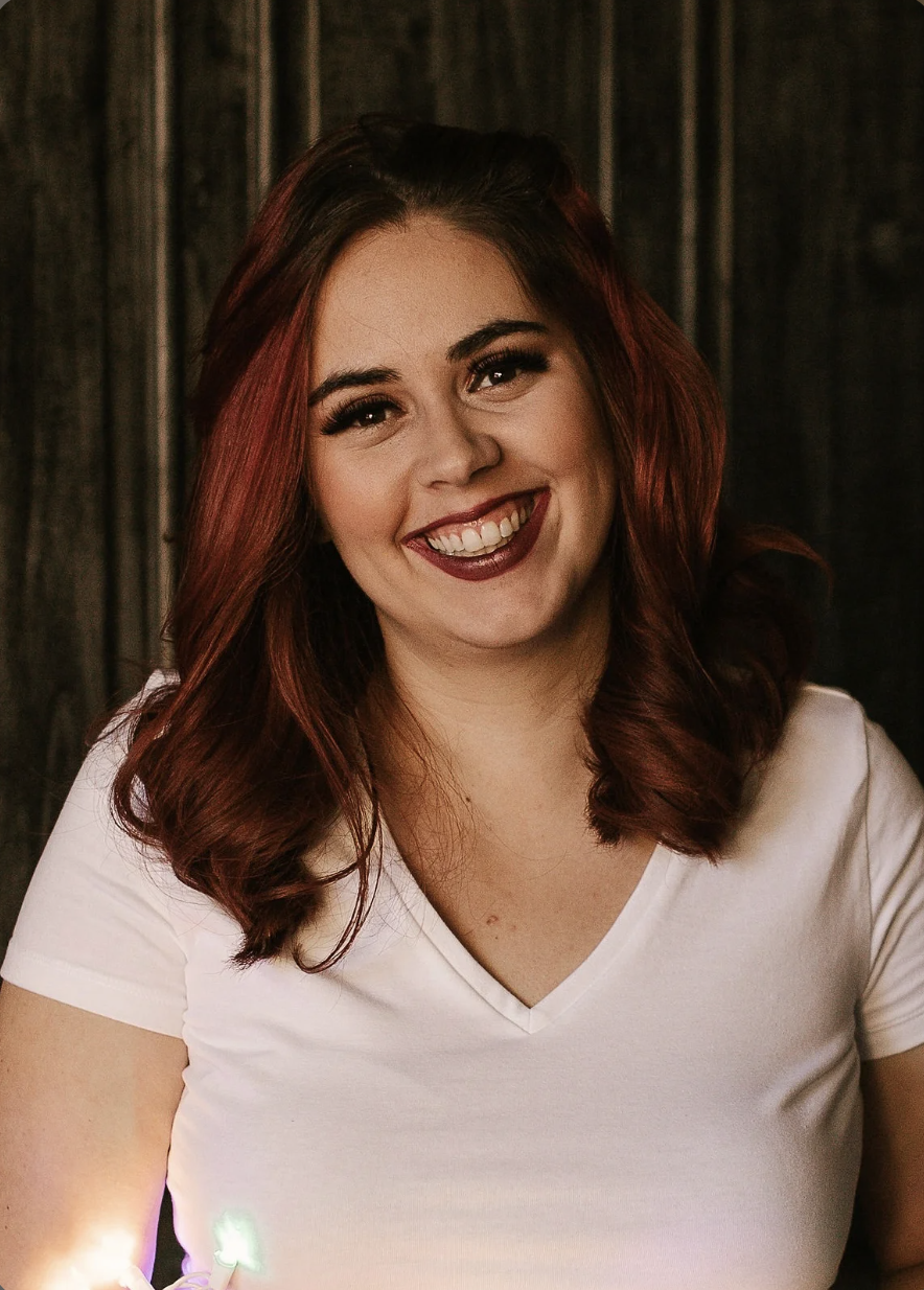 The owner of Cups of Cakes, Sierra Pardo. A smiling woman with red hair and a white V-neck shirt standing in front of a dark wooden background.