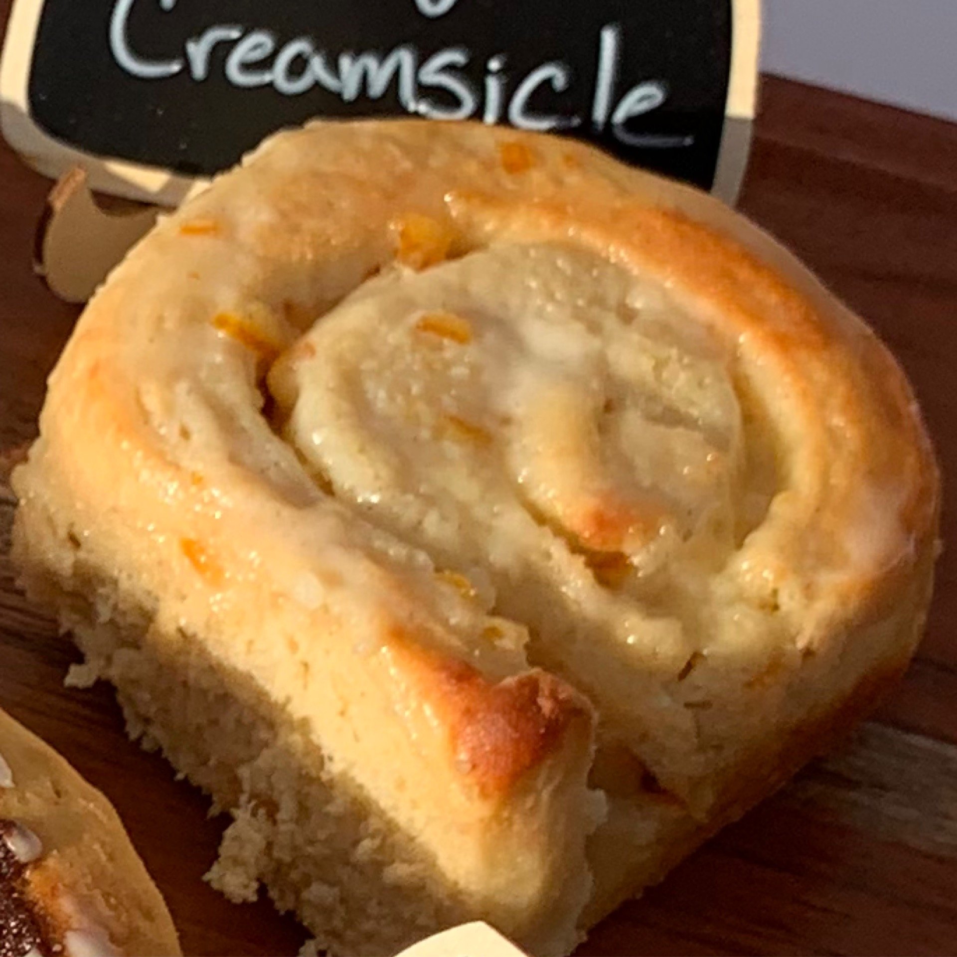 Close-up of a cinnamon roll with icing on a wooden surface, with a small chalkboard sign in the background that reads 'Creamsic'.
