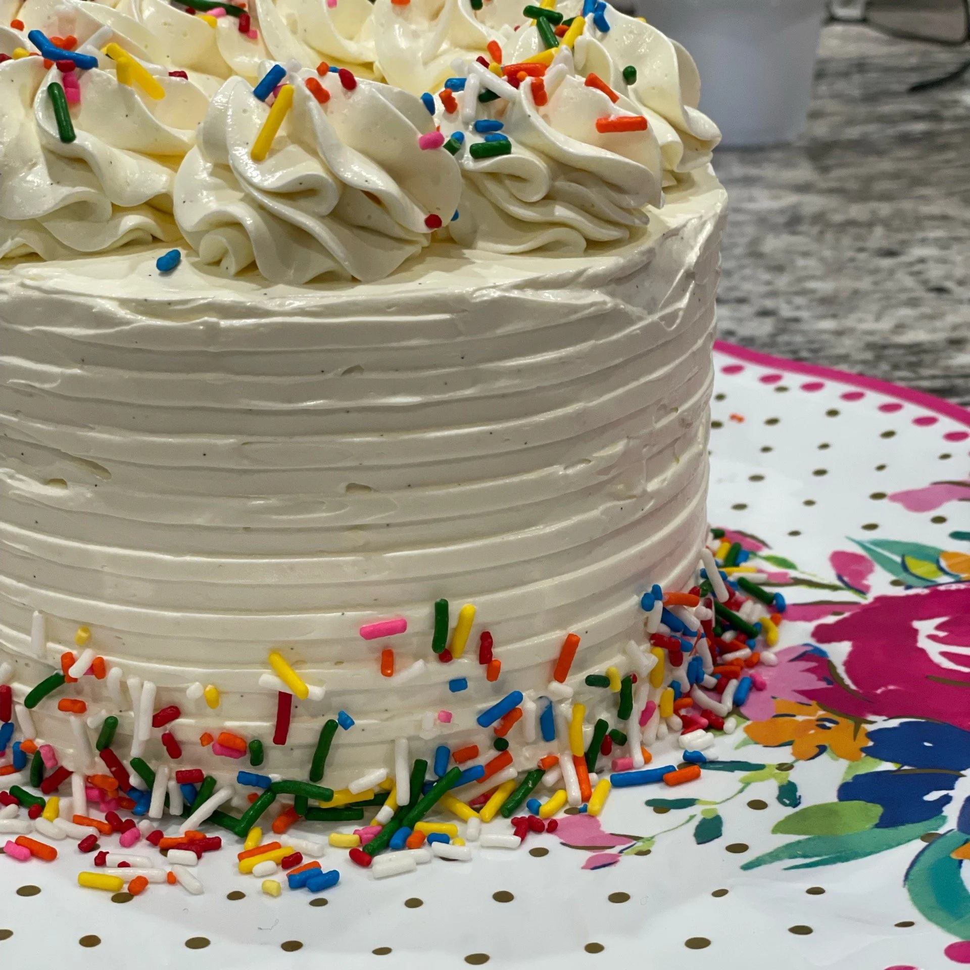 Close-up of a white frosted birthday cake decorated with colorful rainbow sprinkles and white frosting in decorative swirls.