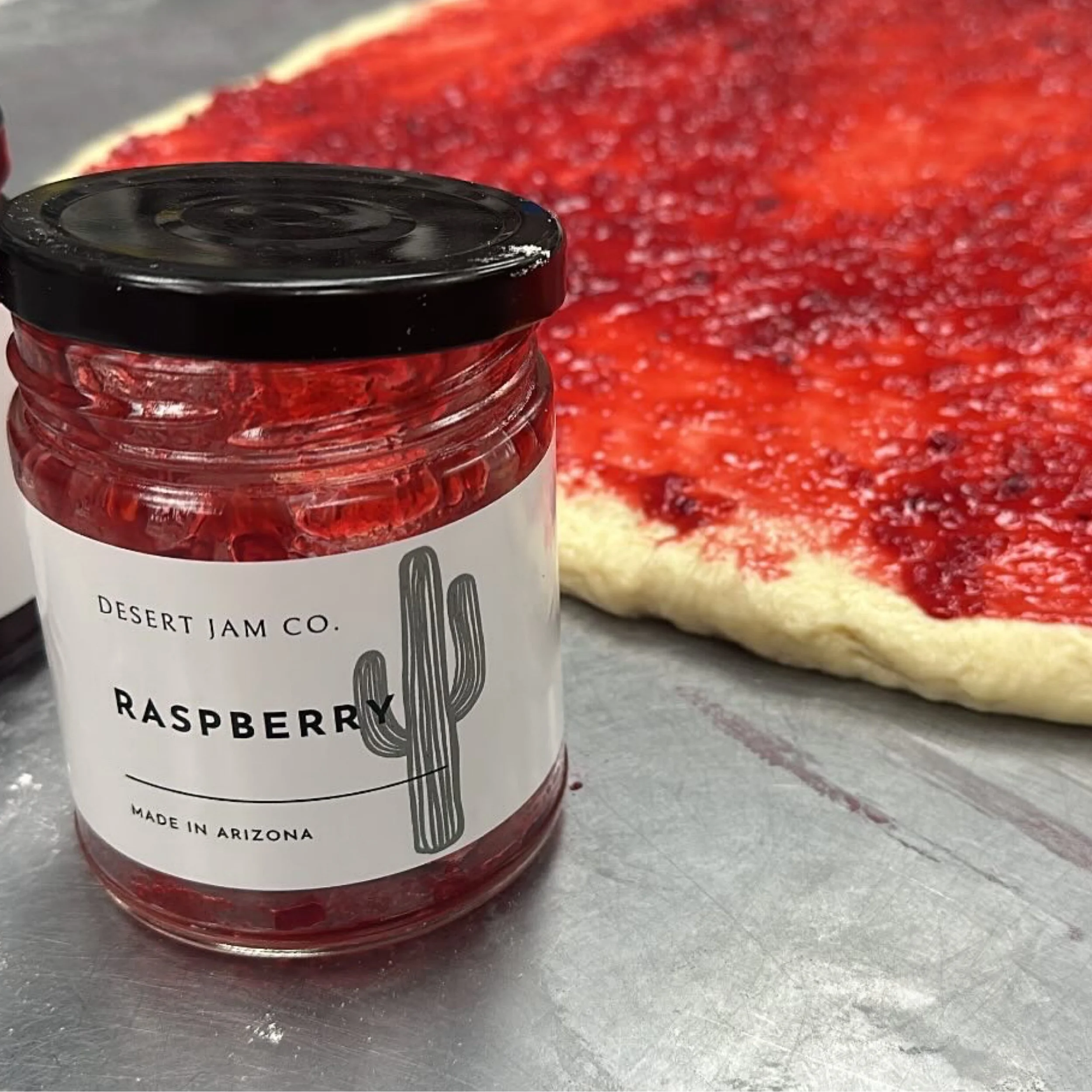 Jar of raspberry jam with a raspberry dessert flatbread in the background.