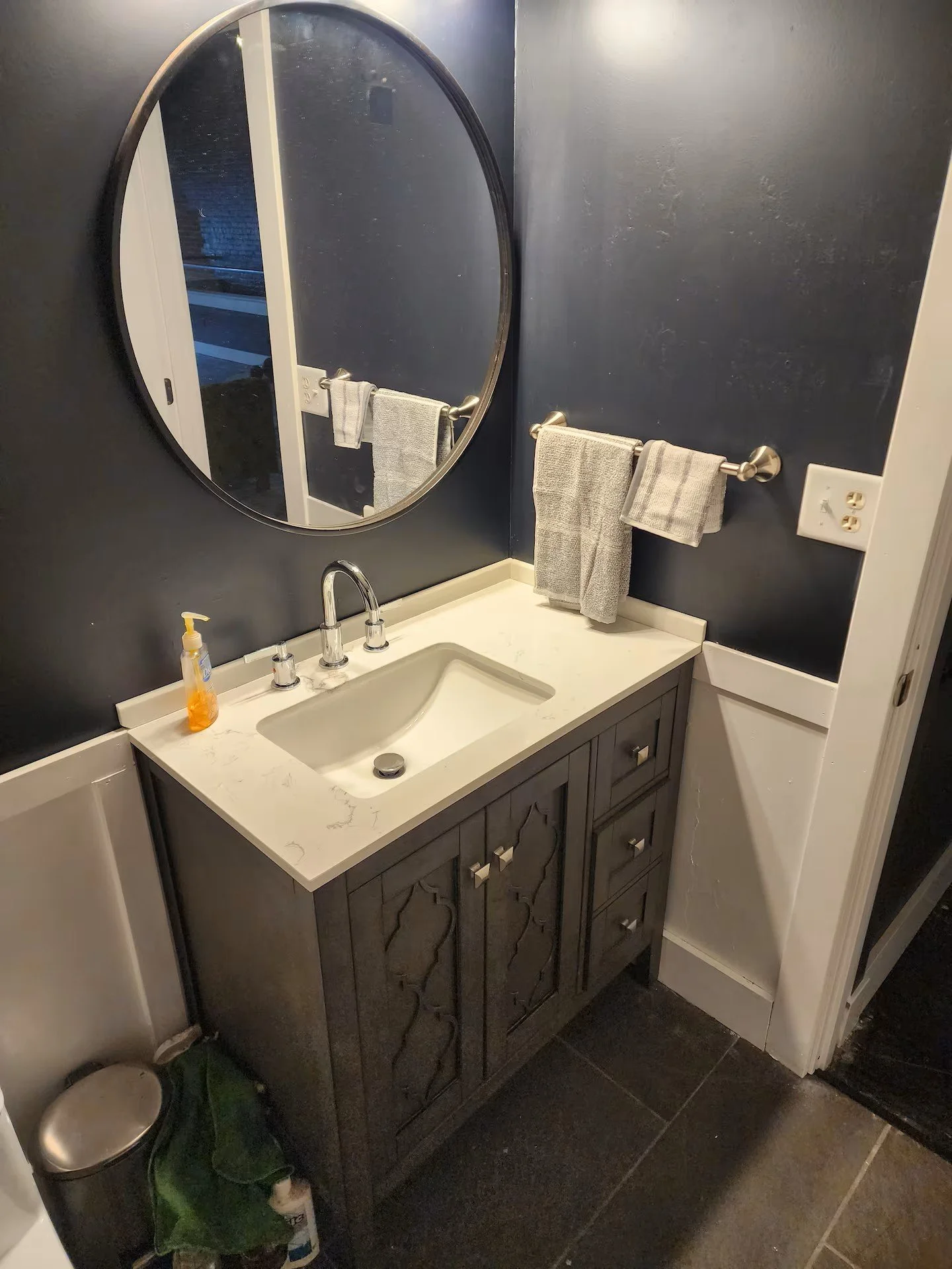 Bathroom vanity with a sink, mirror, towel rack with towels, soap dispenser, trash can, and wall outlets.