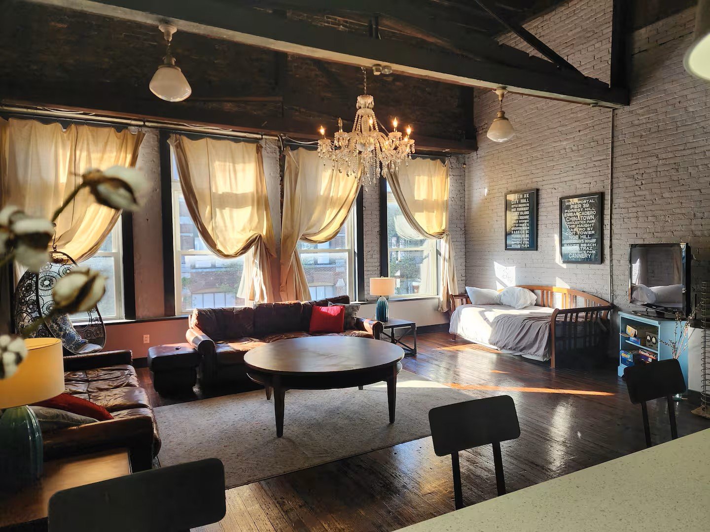 Sunlit urban loft living room with exposed brick walls, large windows draped with cream curtains, vintage leather sofas, a wooden bed, a chandelier, and framed art on the wall.
