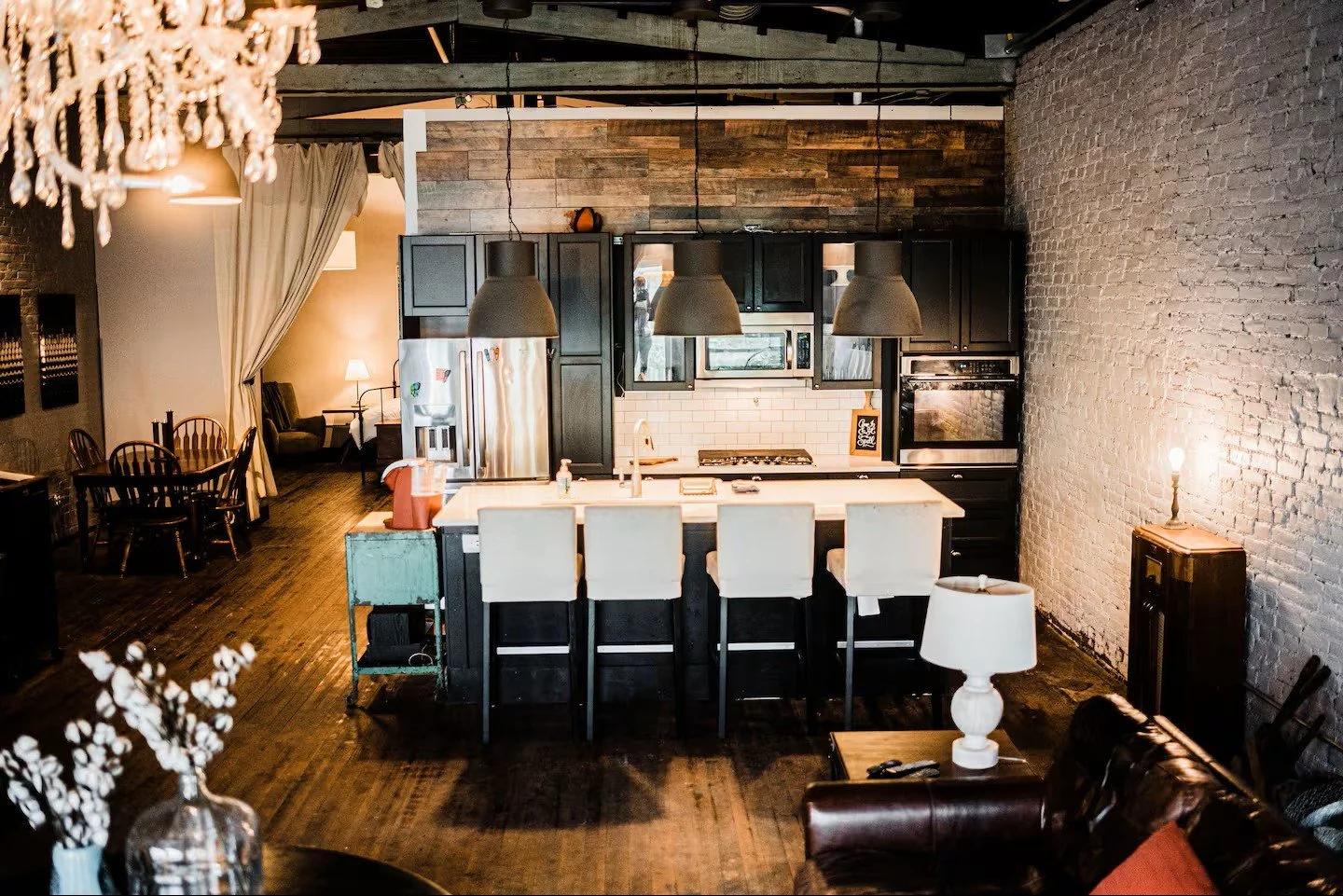 Interior view of a modern kitchen with a white island, black cabinetry, and stainless steel appliances, adjacent to a living area with a lamp, a brown sofa, and a white brick wall.