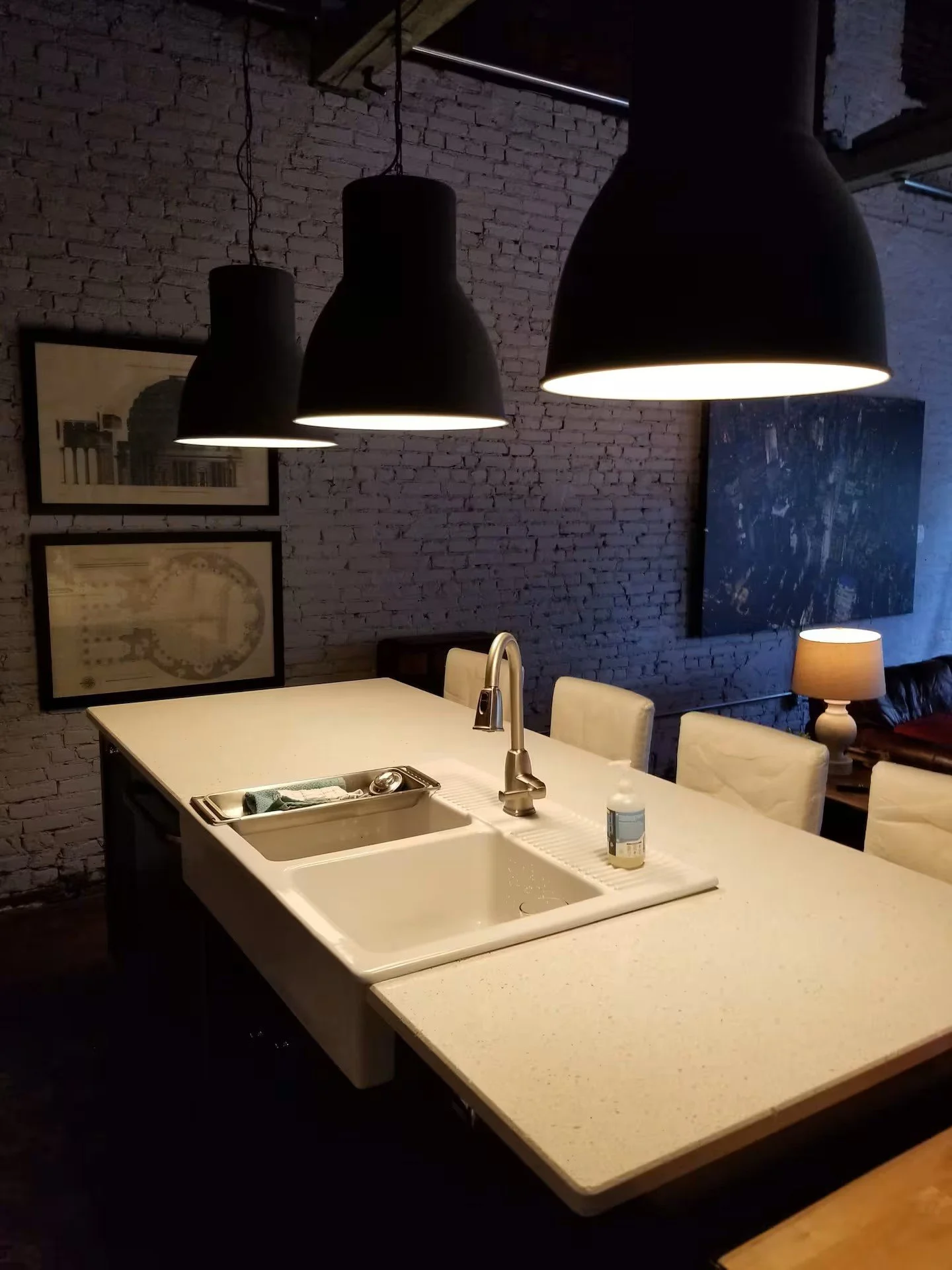 A kitchen with a white countertop, stainless steel sink, black pendant lights, and a brick wall with framed pictures. There is a window, a blue abstract painting, and a table lamp in the background.