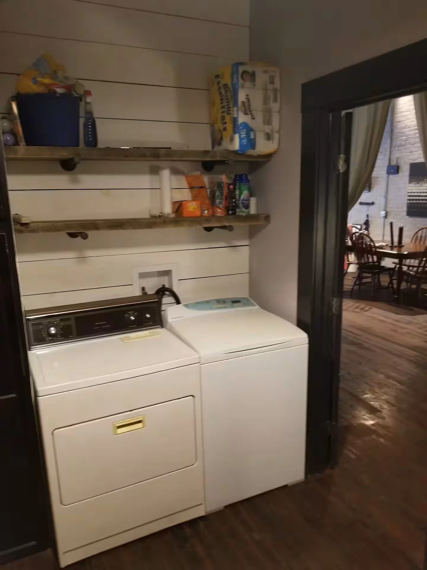 Photo of a laundry area with a washing machine and dryer in front of wooden shelves holding cleaning supplies and paper towels, with a doorway leading to a dining room with wooden chairs and table.