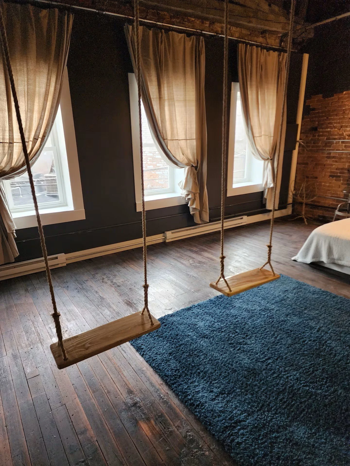 Interior of a room with three windows having beige curtains, exposed brick wall on the right, wooden floor, a blue rug, and a wooden swing hanging from the ceiling in the foreground.