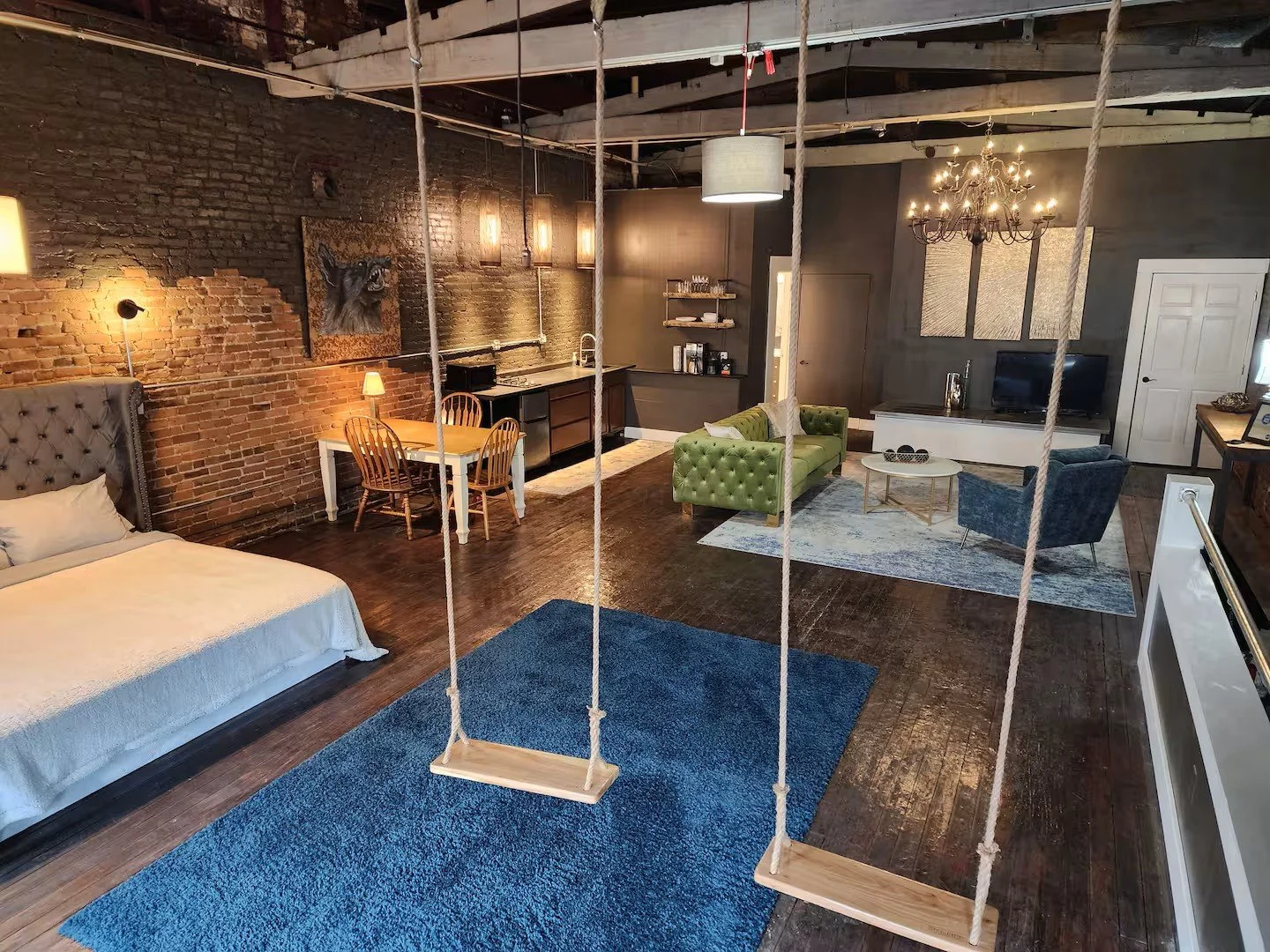A spacious, loft-style apartment with exposed brick walls, hardwood floors, and an open floor plan. The room features a bed with a tufted headboard, a small dining area with a round table and four chairs, a kitchen area with open shelving, a living a