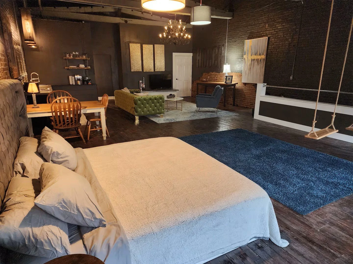 Open-concept living space with a bedroom, dining area, and living room. The bedroom has a bed with beige bedding, a tufted headboard, and pillows. The living room features a green tufted sofa, a gray armchair, a TV on a stand, and a chandelier. A swi