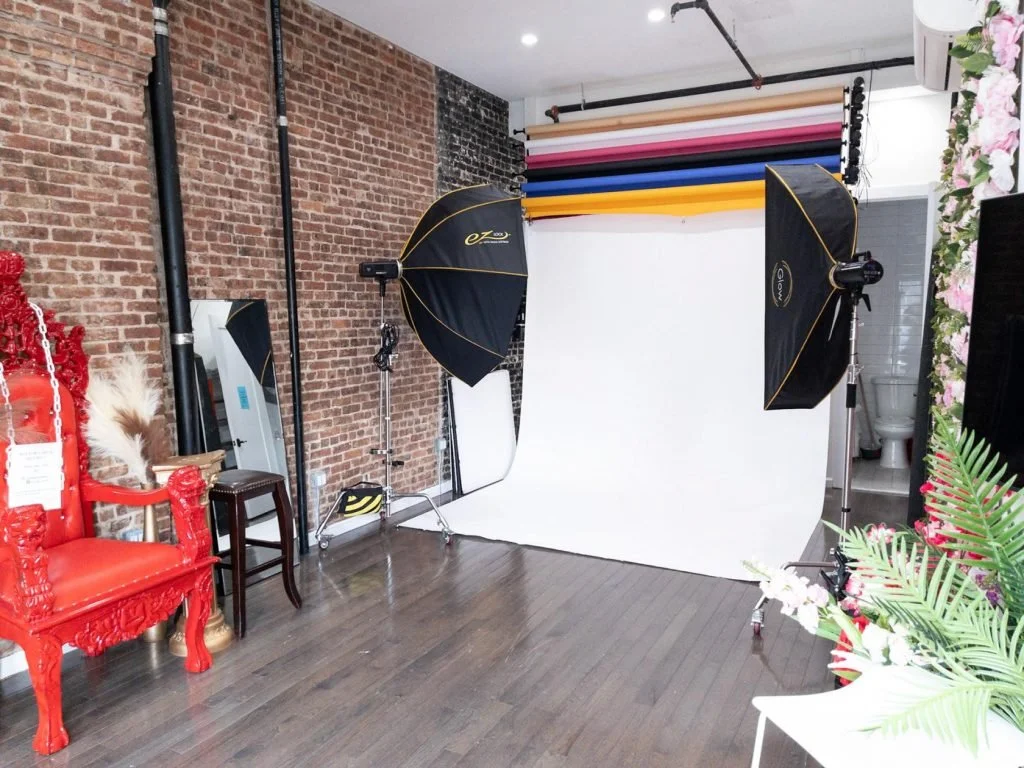 Photography studio setup with a white backdrop, softbox lights, and colored backdrop rolls, with a decorative red chair and floral accents.