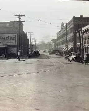 A historic black and white photograph of a small town main street with vintage cars parked along the curb, old brick buildings, and utility poles with power lines.