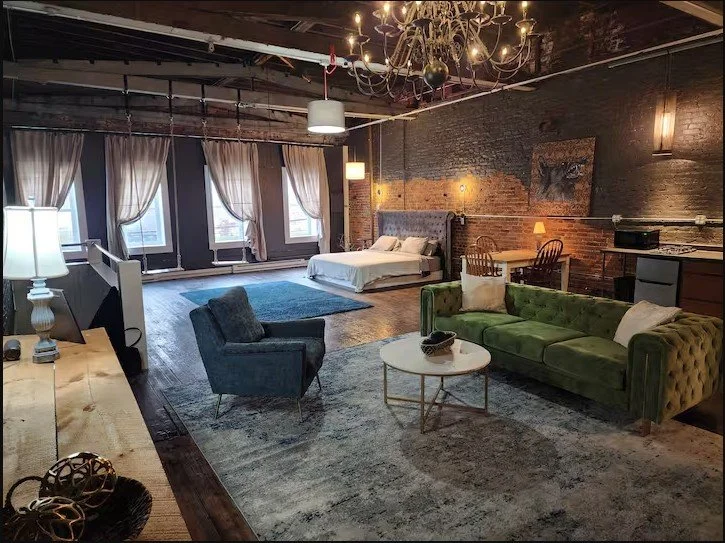 Living space with exposed brick walls, large windows with curtains, and a mix of modern and vintage furniture including a green velvet sofa, a gray armchair, a white bed, a chandelier, and a blue area rug.