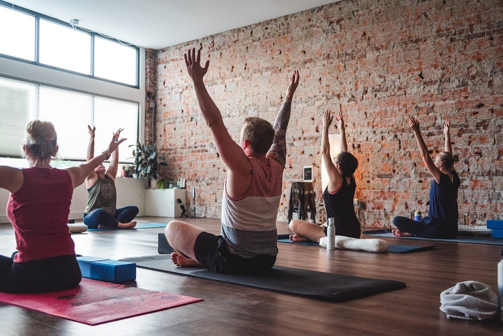 A group of people participating in a yoga class in a room with a brick wall, sunlight coming through windows, and yoga mats. They are sitting cross-legged with arms raised.