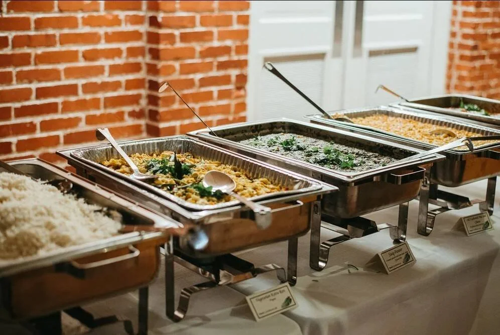 Buffet table with trays of rice, pasta, and other hot dishes against a brick wall.