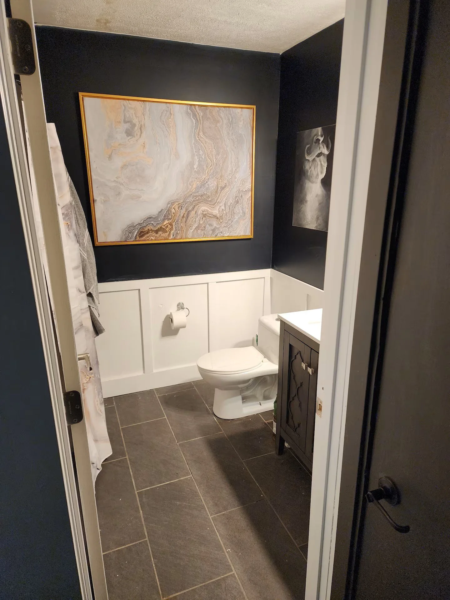 A bathroom with dark walls, a large abstract painting with a gold frame, a smaller black and white portrait of a dog, a toilet, a dark wood cabinet, and a gray tiled floor.