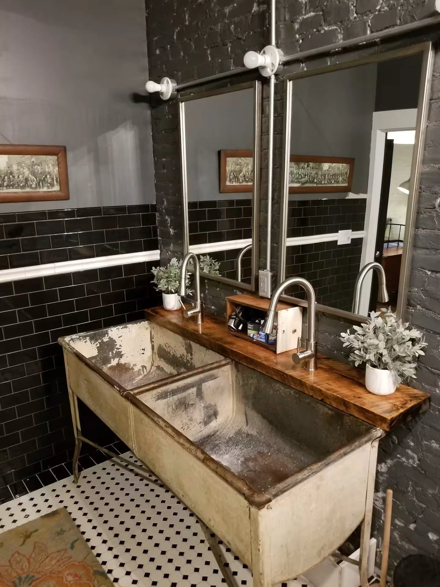 A vintage metal bathtub with peeling paint and rust, positioned underneath a wooden countertop with two modern silver sinks and faucets. Above the countertop are two large rectangular mirrors framed in metal, mounted on a brick wall painted gray. The
