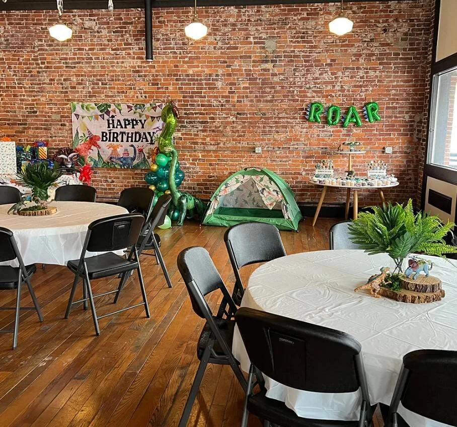 Indoor birthday party setup with round tables covered with white tablecloths, black chairs, a brick wall decorated with birthday banners, balloons, and a small green tent. There are also green letter balloons spelling 'ROAR' and a table with food and desserts.