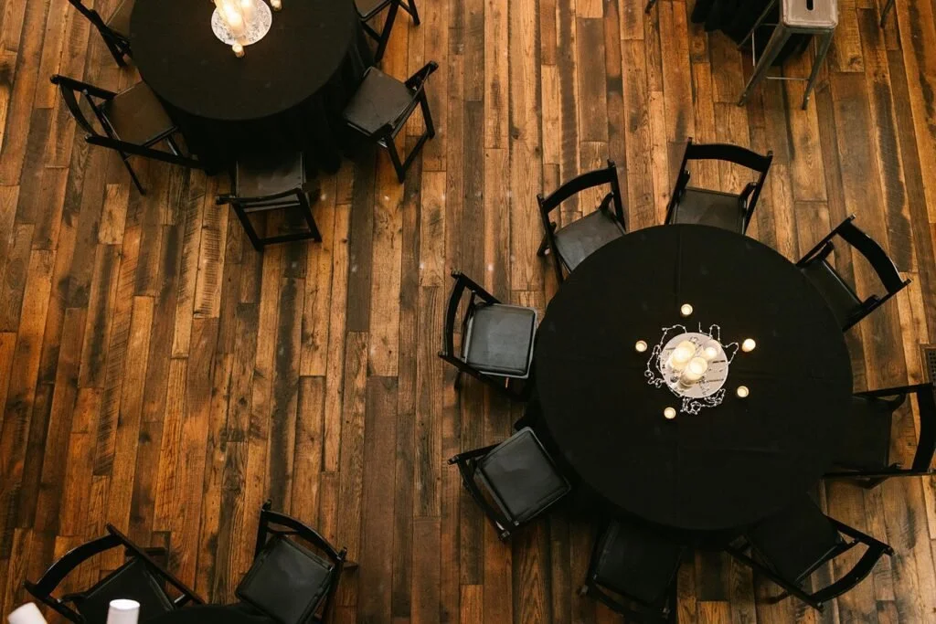 Two round tables with black tablecloths and black chairs around them are set up on a wooden floor in a room, with a chandelier hanging over each table.