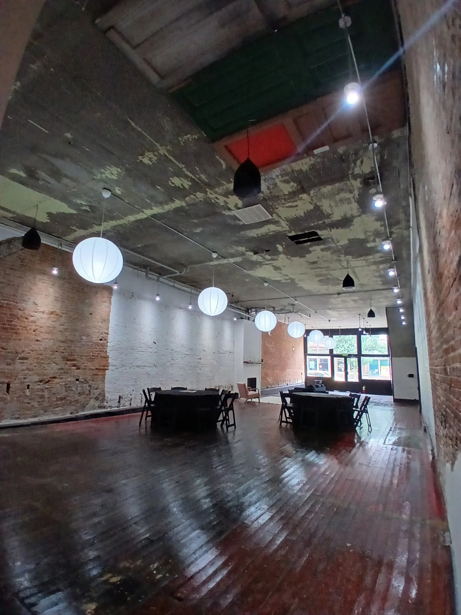 An industrial-style interior space with exposed brick walls, a weathered wooden ceiling, and hanging paper lantern and black pendant light fixtures. There are round tables with chairs on a polished wooden floor, large windows letting in natural light