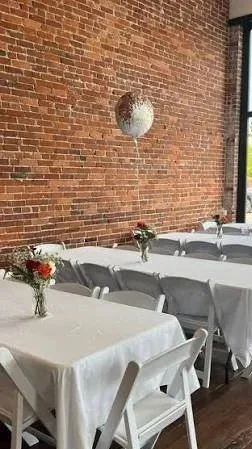 Wedding reception setup with long tables covered in white tablecloths, small flower arrangements, brick wall, and a hanging spherical light fixture.