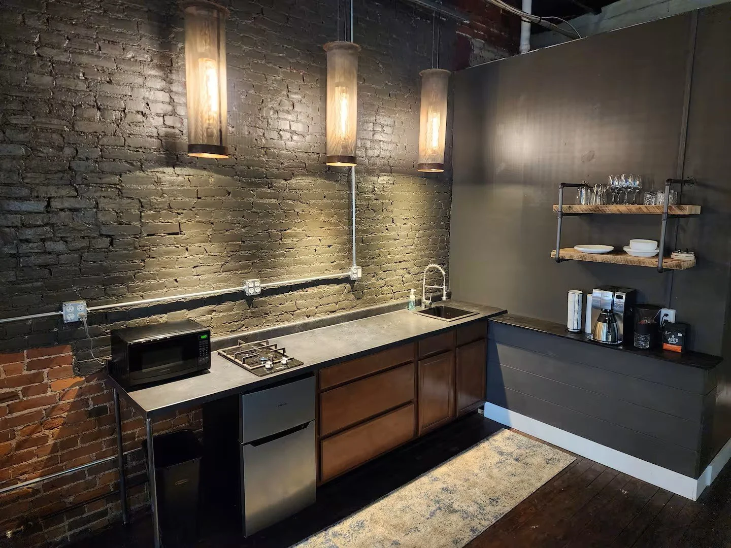 Small kitchen with black brick walls, exposed piping, hanging cylindrical light fixtures, wooden cabinets, a mini fridge, microwave, small sink, coffee maker, glasses, plates, and bowls on floating shelves