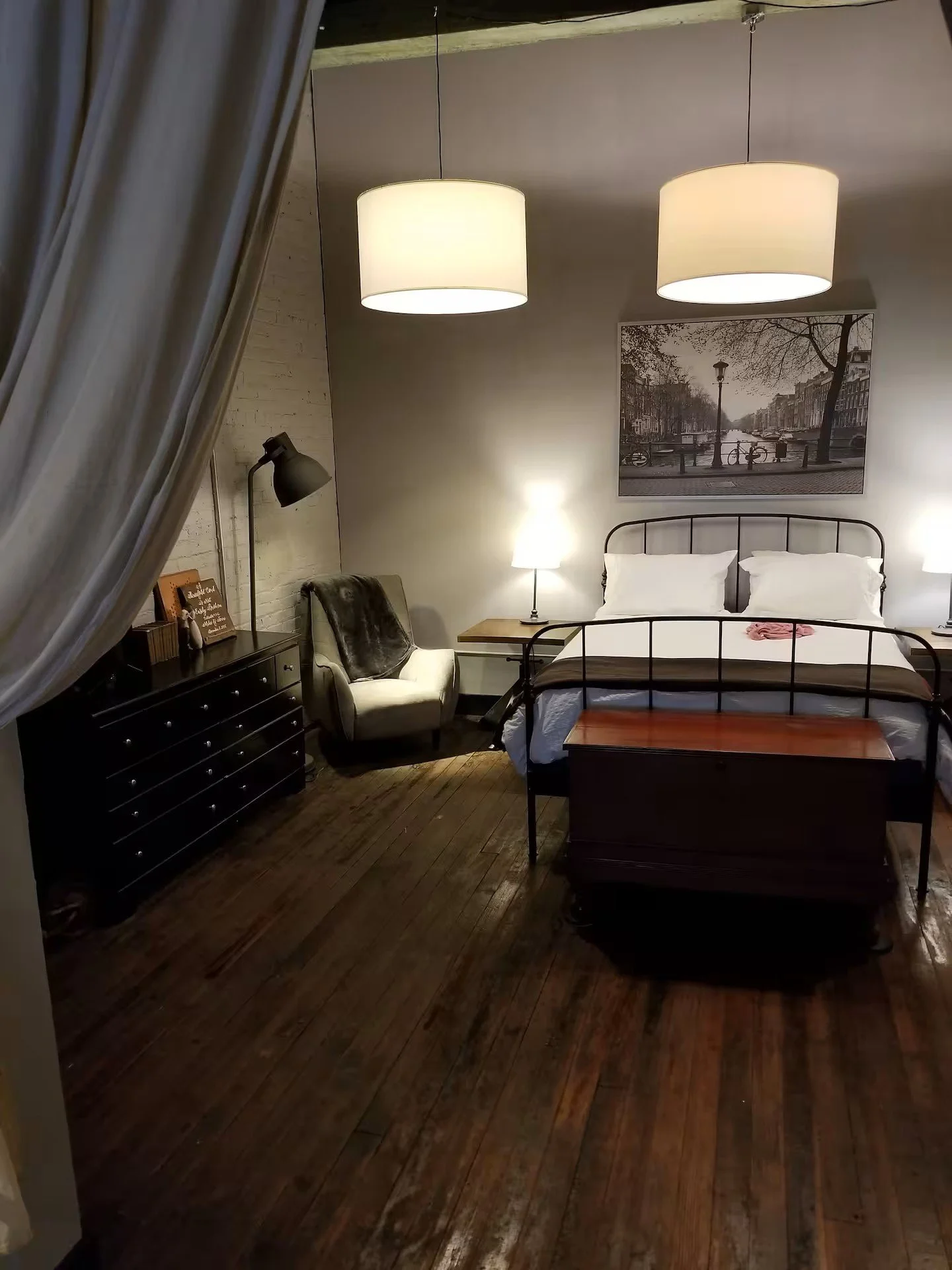 Bedroom with white brick wall, bed with white pillows and pink towel, wooden floor, black metal bed frame, black dresser with picture and decor, vintage armchair with throw pillow, two bedside lamps, black floor lamp, and framed cityscape photo on wa