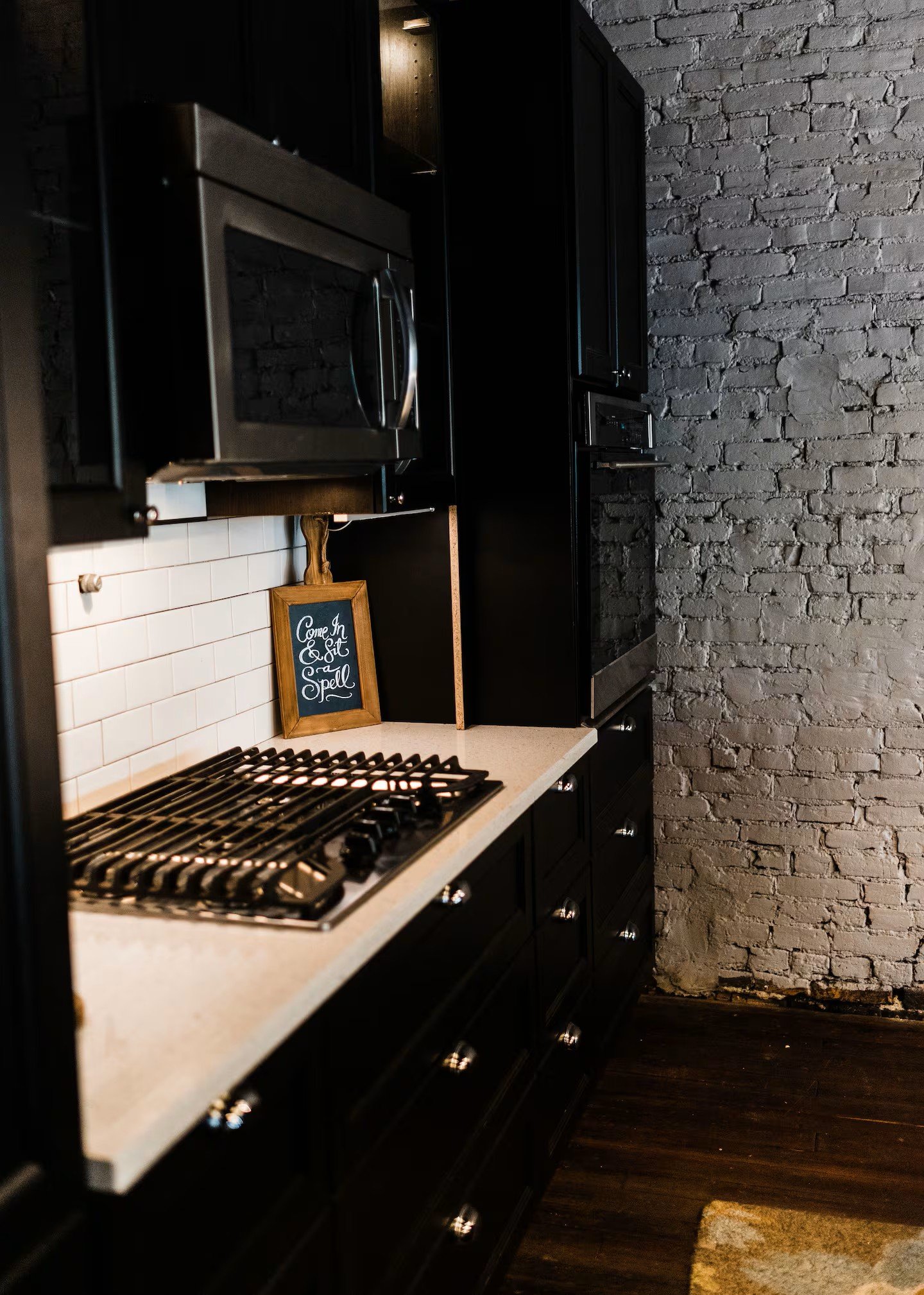 Close-up of a modern kitchen with black cabinets, a beige countertop, a gas stovetop, a small chalkboard sign that reads 'Come In & Sit A While,' and a brick wall in the background.