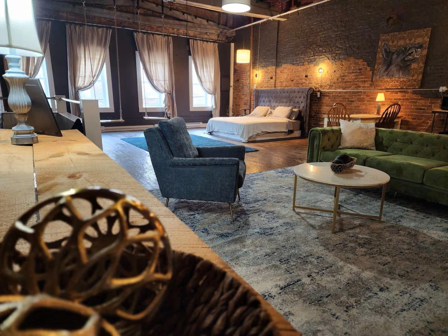 A spacious living area with a cozy bedroom setup, exposed brick wall, large windows with curtains, a blue armchair, a green sofa, a circular coffee table, and a bed with a tufted headboard, decorated with artwork and lamps.