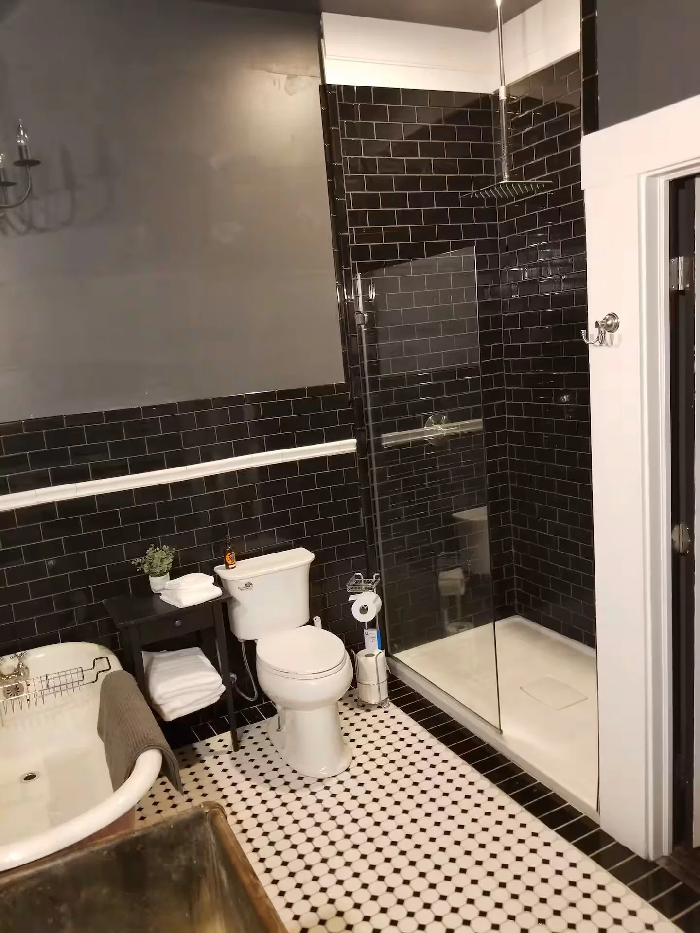 A bathroom with black subway tile walls, a glass shower enclosure with a rainfall showerhead, a toilet, a small black side table with towels and a plant, and an original black and white hexagonal tile floor.
