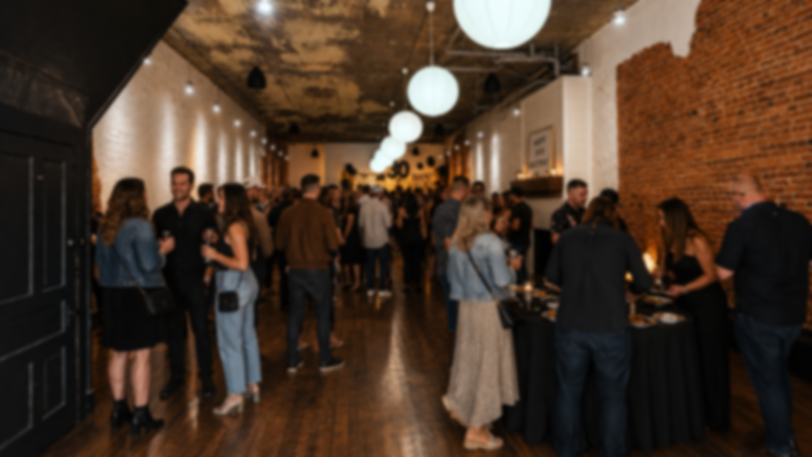 People socializing and dining at a party or event in a modern, industrial-style venue with brick walls and hanging white globe lights.