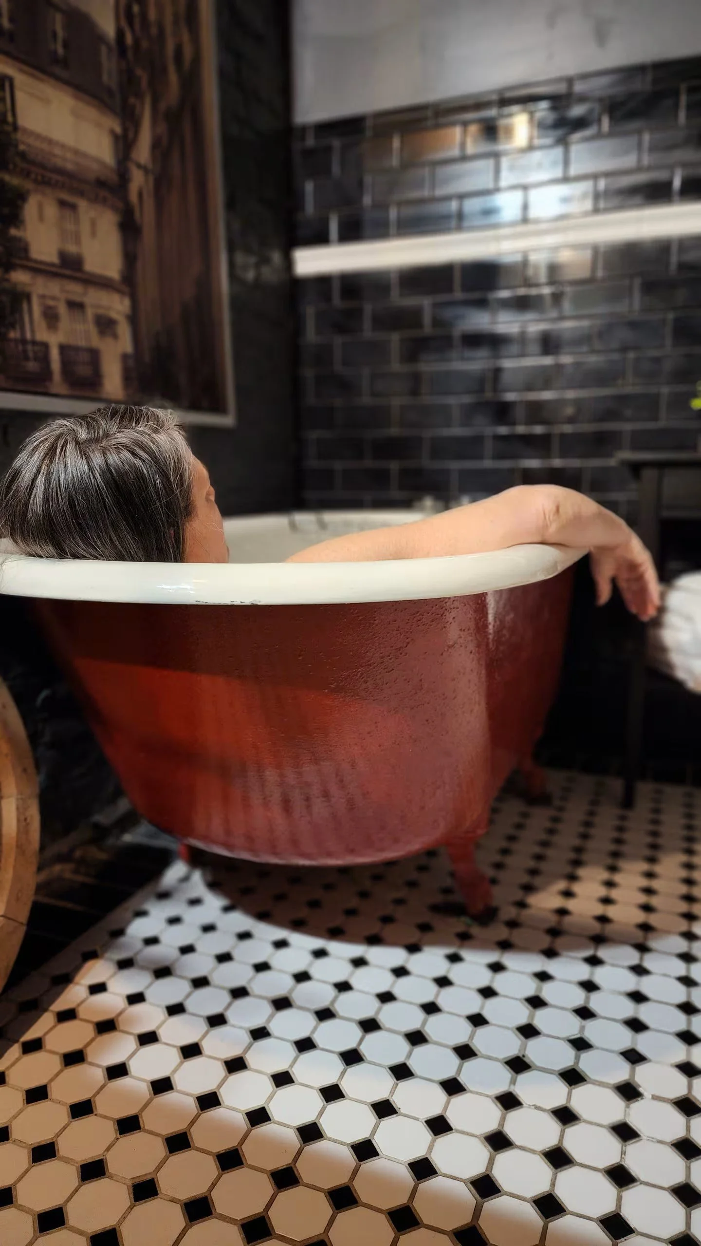 Person relaxing in a vintage red bathtub with a white interior, situated on a black and white hexagon tile floor, with a dark brick wall and framed artwork in the background.