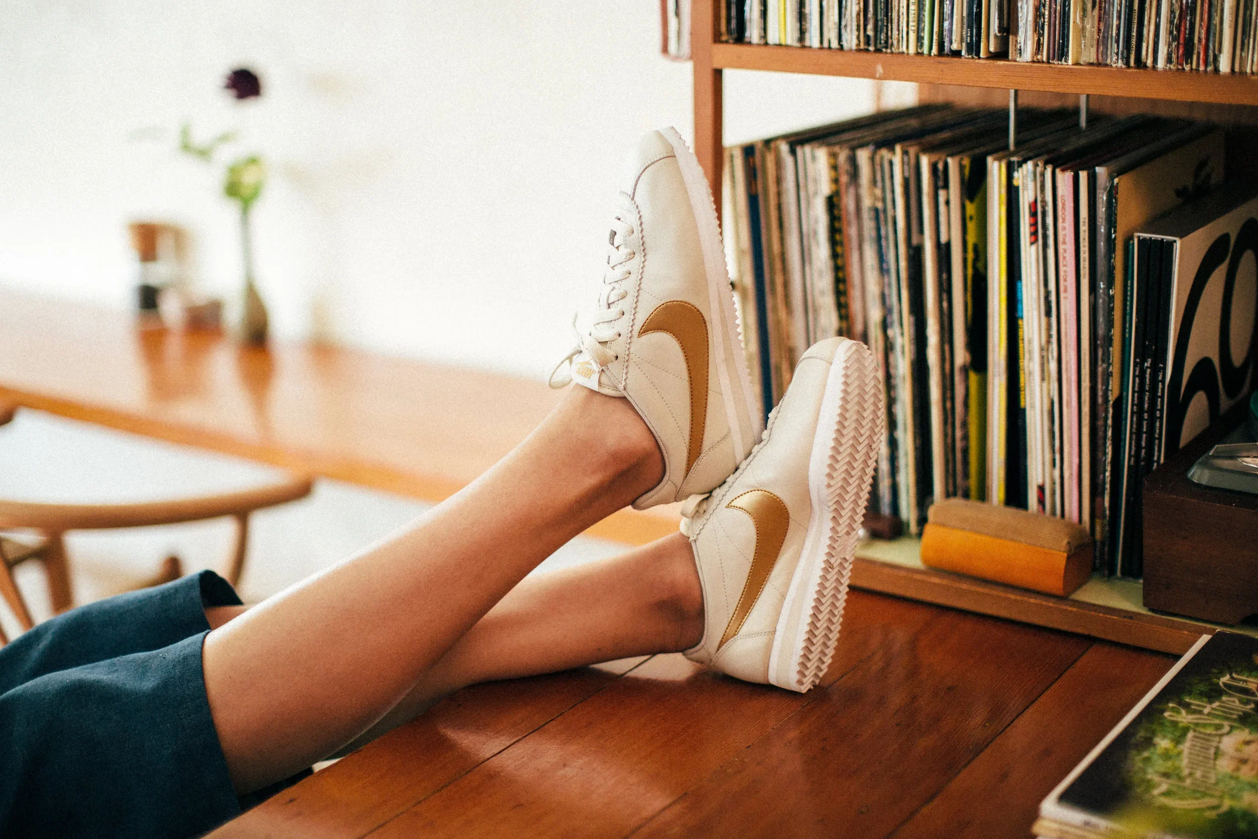 Person relaxing with feet up on a wooden table, wearing white and brown sneakers, next to a bookshelf filled with vinyl records.
