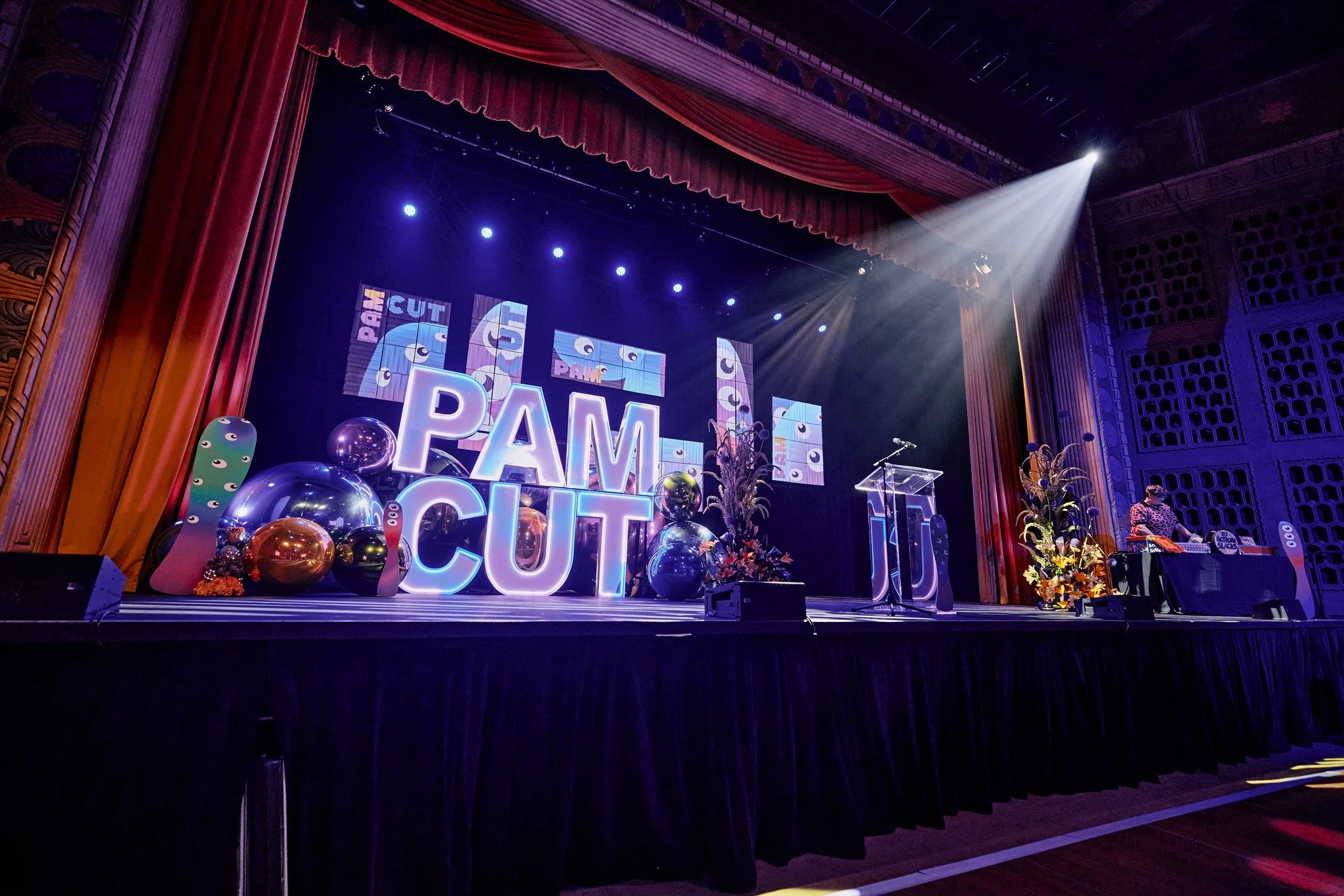 Stage decorated with large illuminated letters spelling 'PAM' and 'CIT', colorful balloons, and flowers with a red curtain backdrop and stage lights.