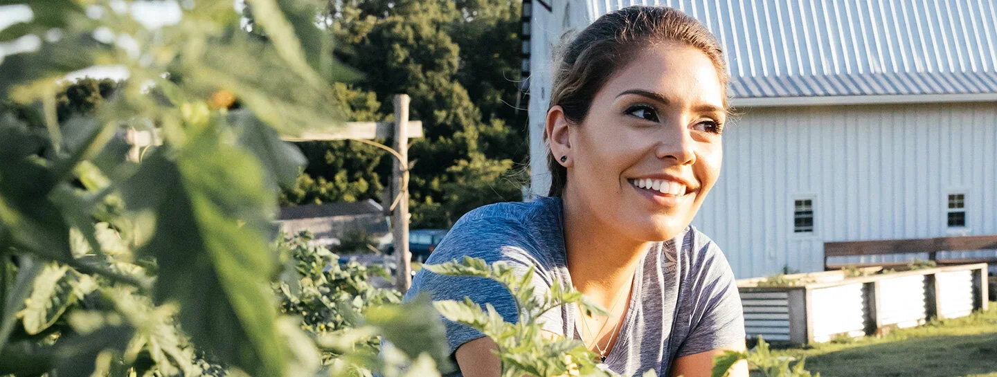 A woman with brown hair smiling outdoors in front of a white barn, surrounded by green plants.