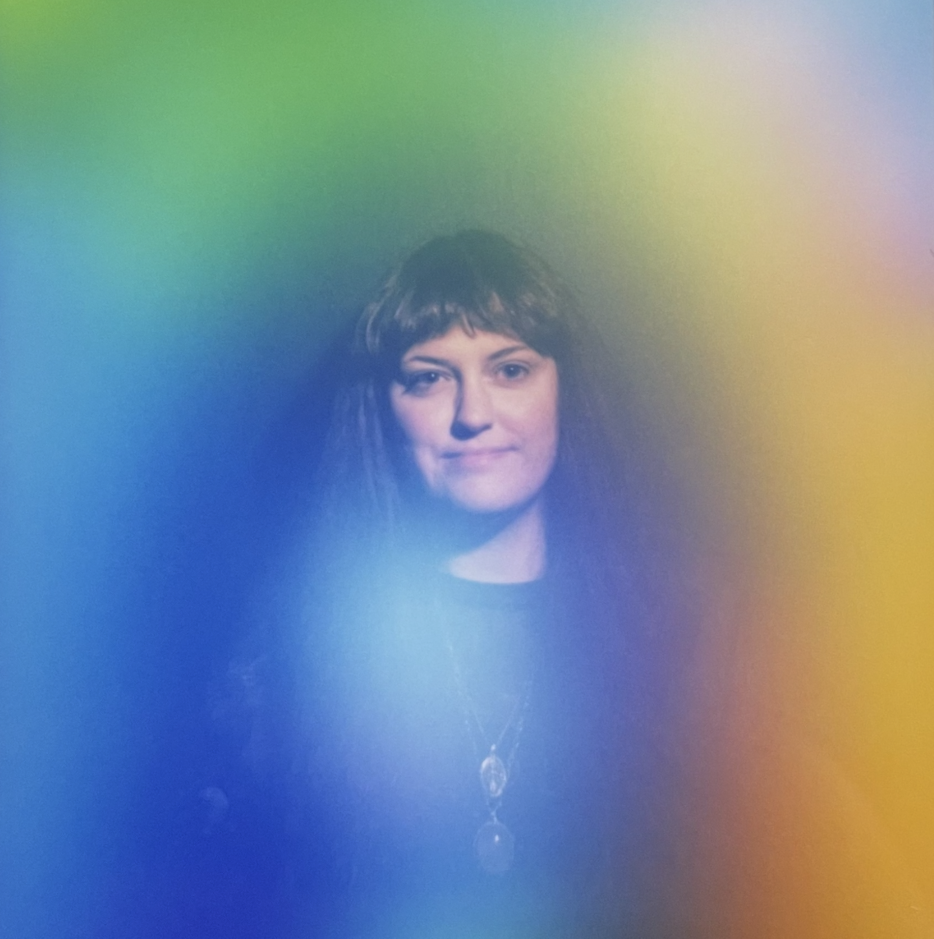 A person with short dark hair and light skin looking directly at the camera, with rainbow-colored light effects in the background.