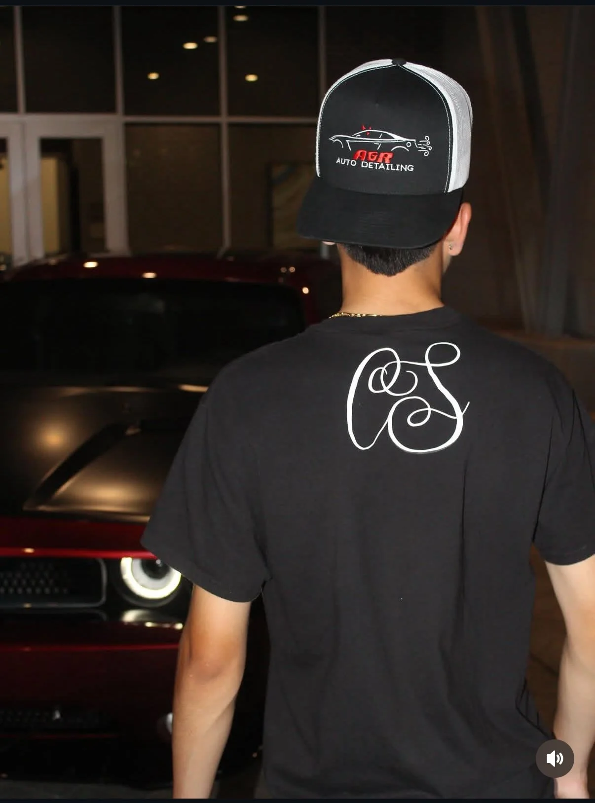 Person standing with back to camera, wearing black T-shirt with white script design on back and black and white trucker cap with 'AG Auto Detailing' logo, in front of a red vehicle inside a building.