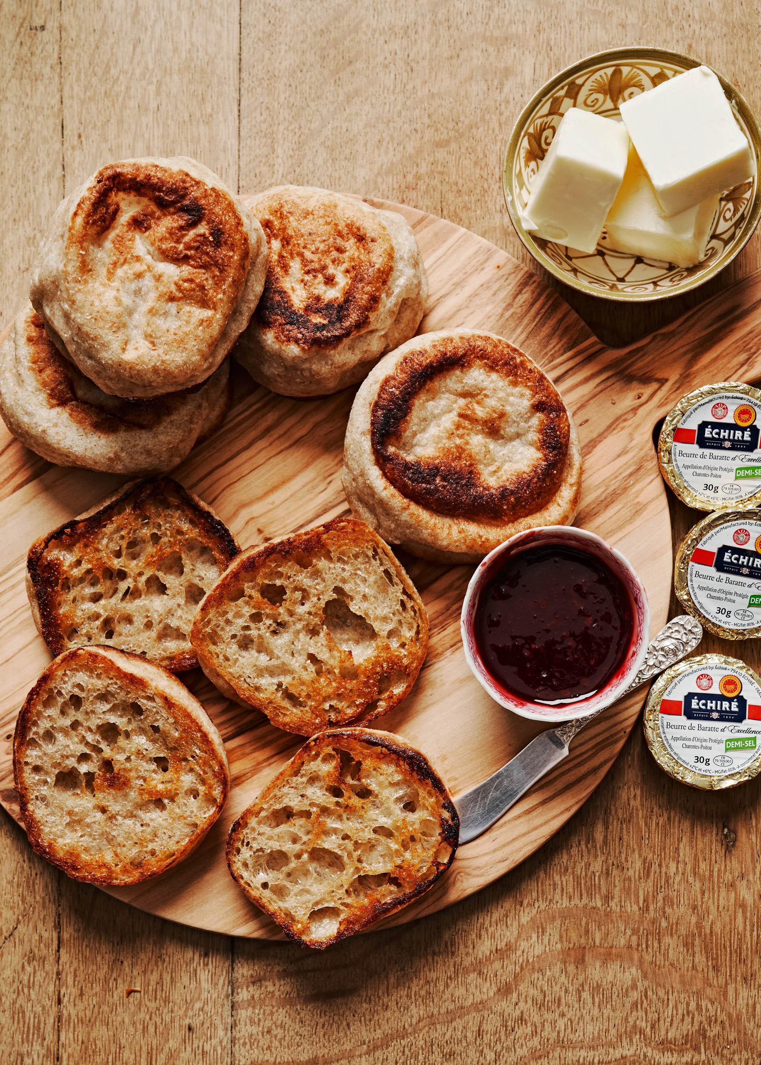 Sliced toasted bread on a wooden serving board, a bowl of butter, a jar of jam, and a knife, on a wooden table.
