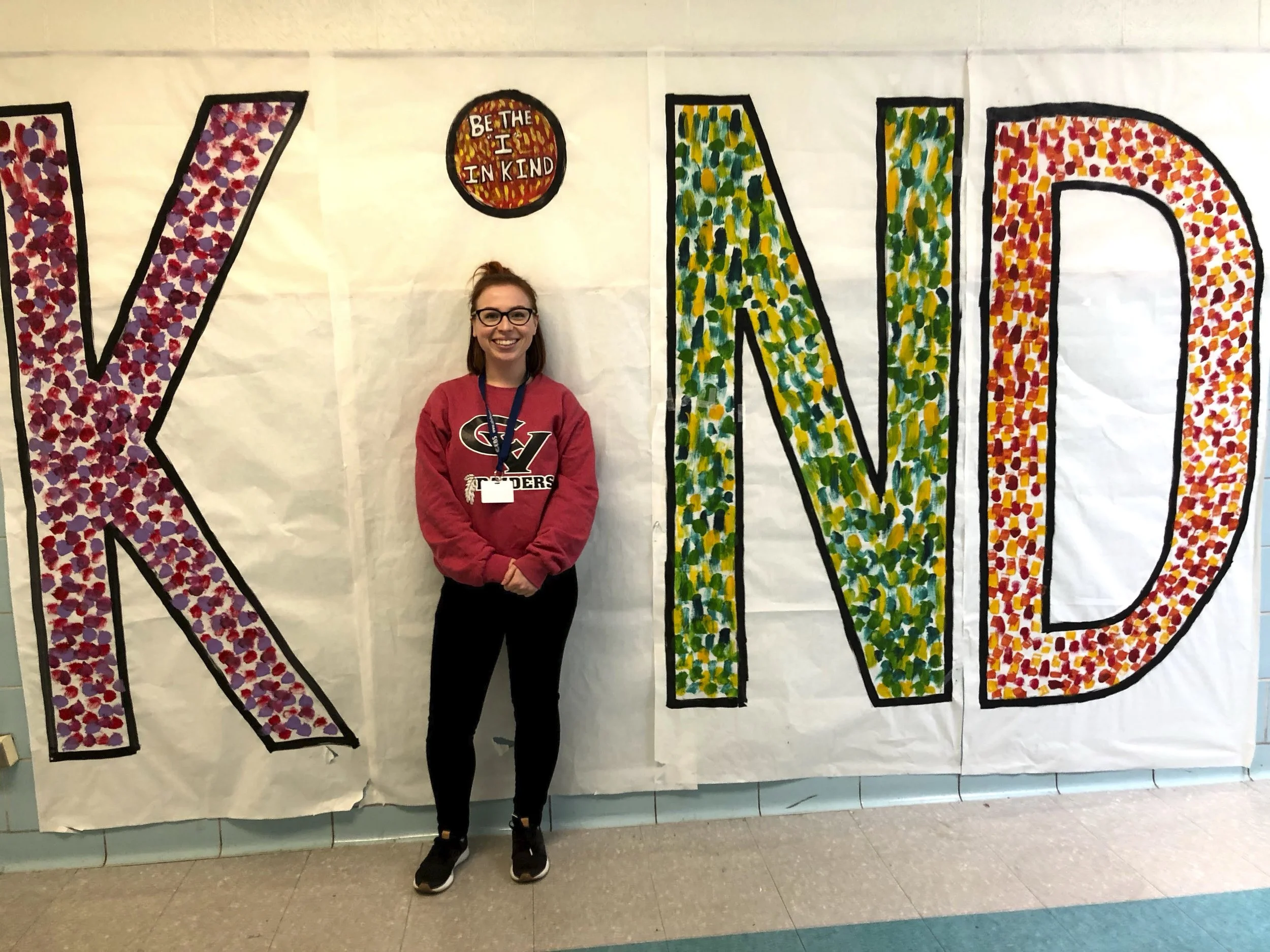 A woman with glasses and a red sweatshirt standing in front of a decorated wall with large text that spells 'KIND.' "Be the I in kind" is written inside a circle on the wall.