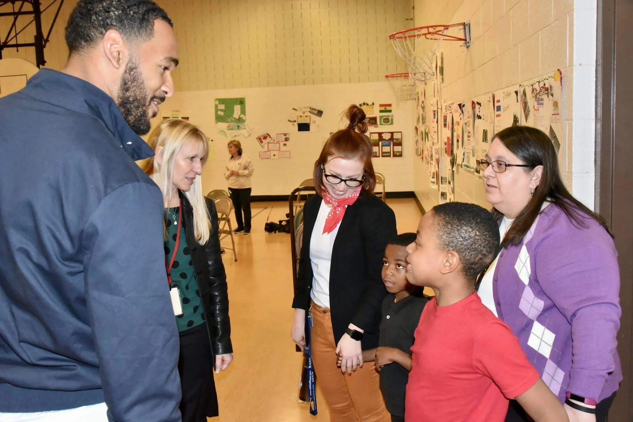 Group of people, including children and adults, engaged in conversation inside a community center decorated with school projects on the walls.
