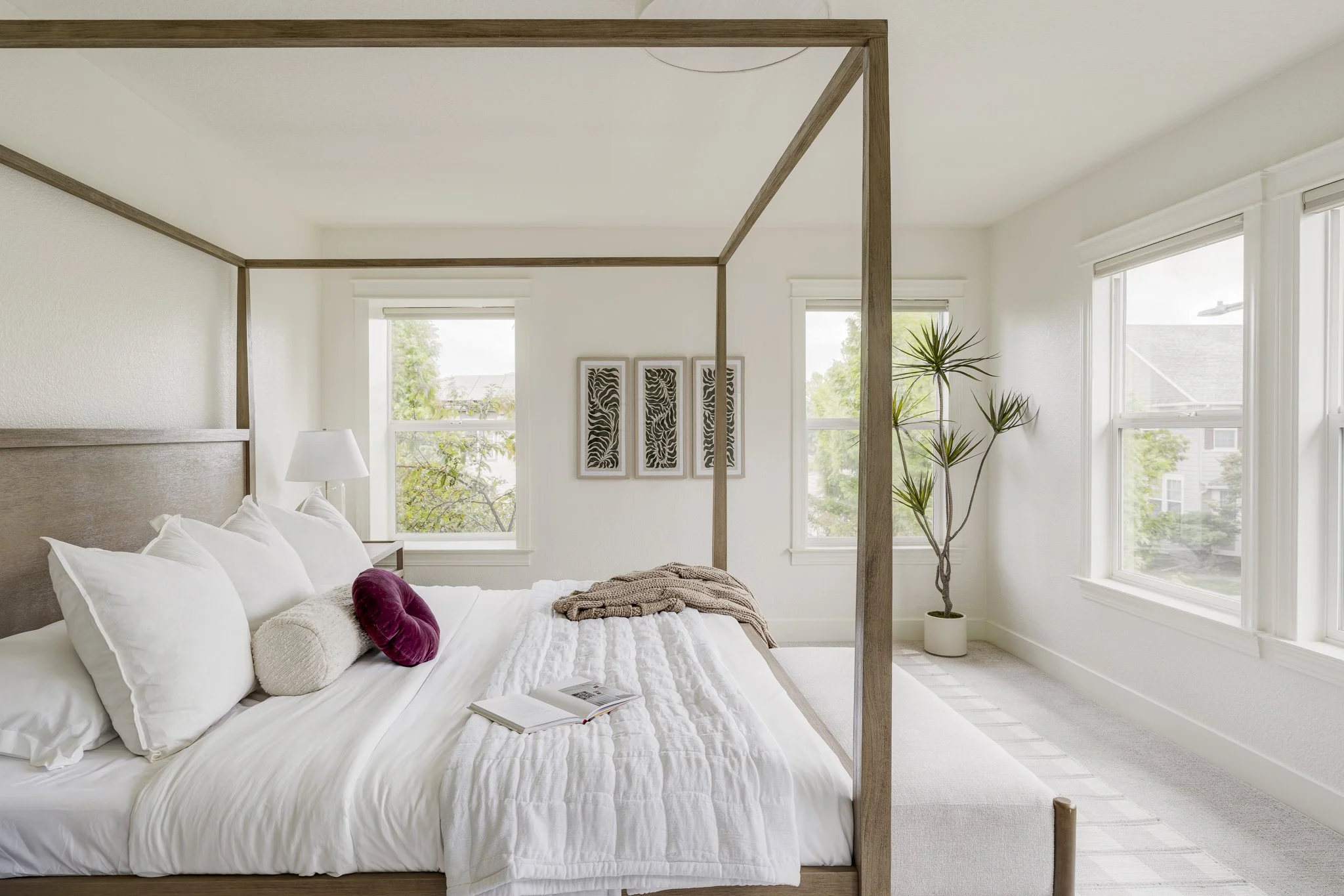 A bedroom with a four-poster wooden bed, white bedding, and pillows, including a round purple pillow and a gray bolster. There is a bedside table with a white lamp, three framed decorative art pieces on the wall, a potted plant near the windows, and three large windows with white blinds allowing natural light to fill the room.