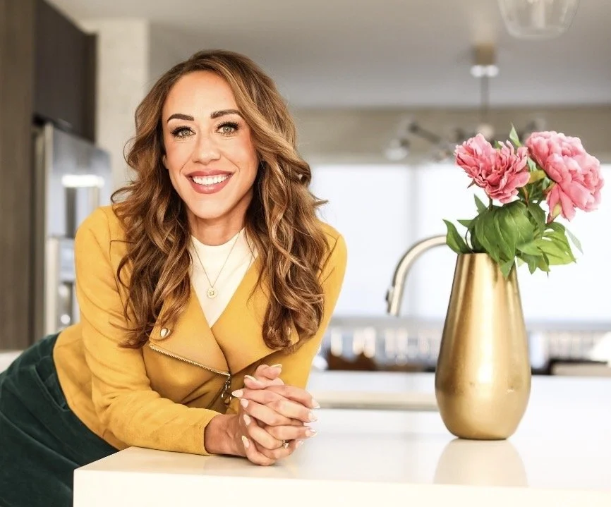 A woman with wavy brown hair, wearing a yellow jacket and smiling, leaning on a kitchen counter.
