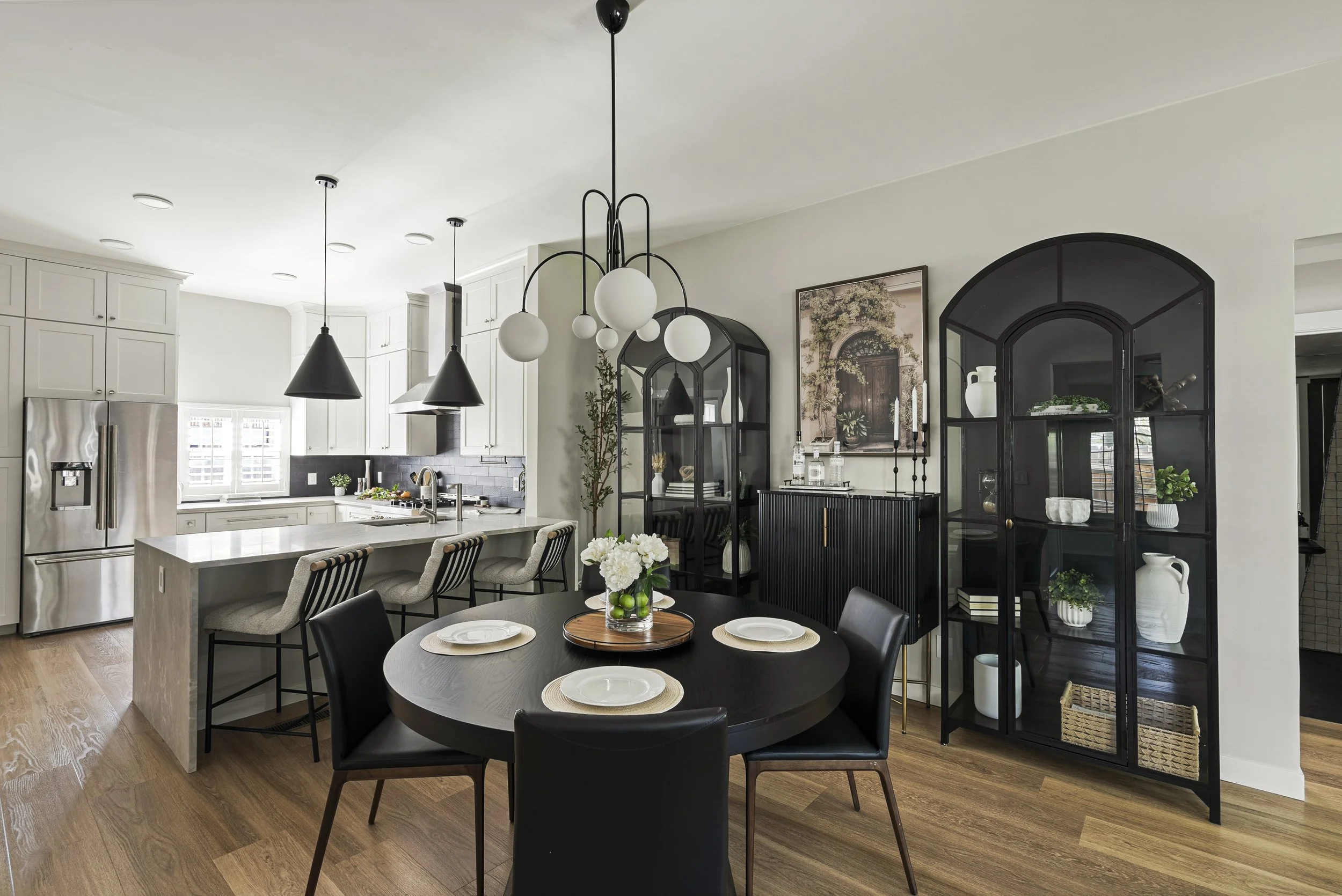Modern dining room and kitchen with black round table and six black chairs, black glass display cabinet with decorative items, white kitchen cabinets, stainless steel refrigerator, and contemporary lighting fixtures.