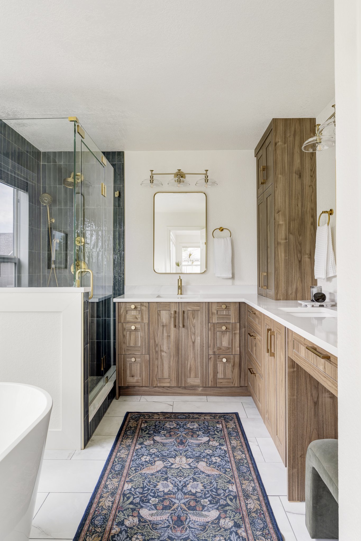 A bathroom with a wooden vanity, a mirror, a towel rack with a white towel, a glass shower with dark tiles, and a decorative rug on white tile floor.