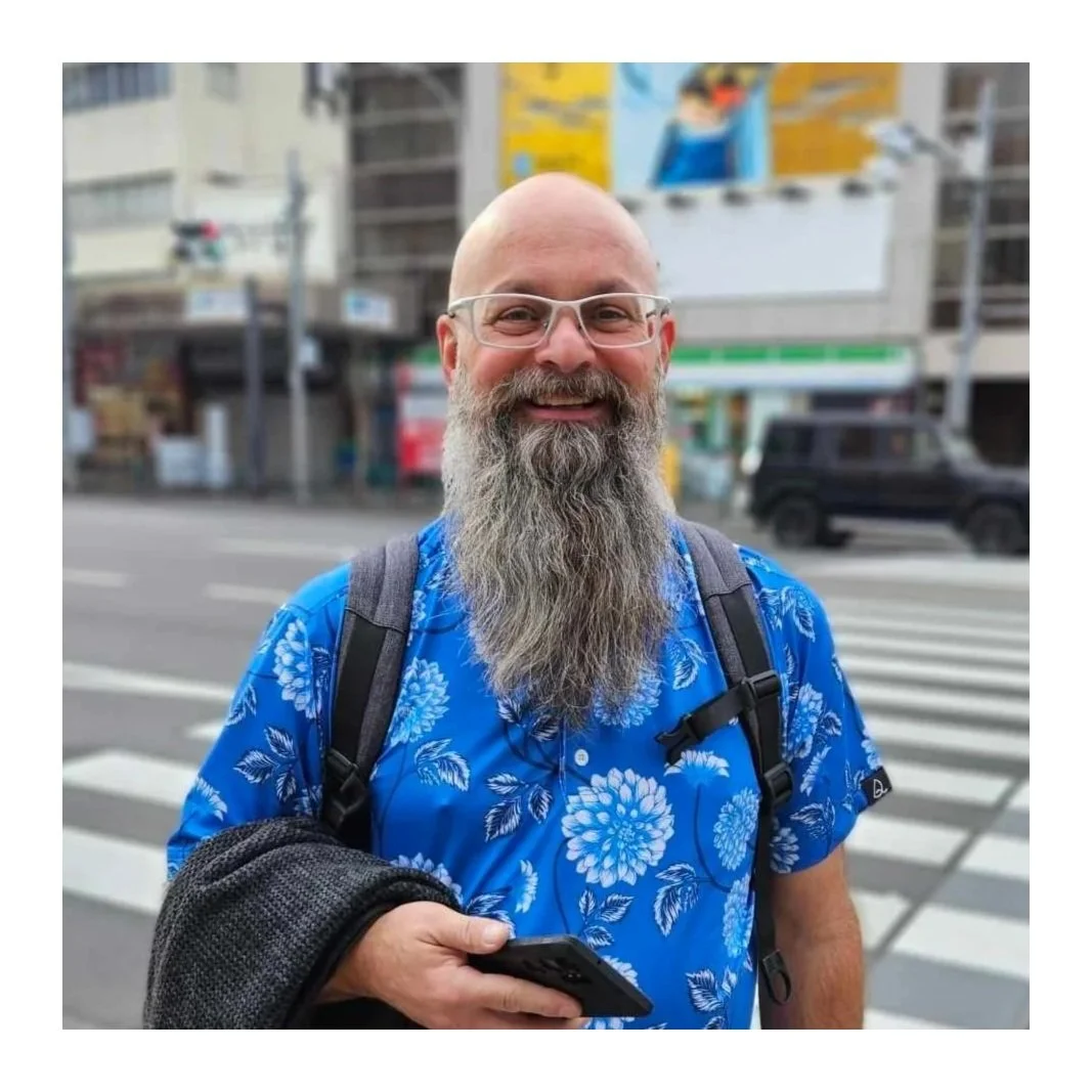 A smiling man with a long gray beard, wearing glasses and a blue floral shirt, standing on a city street at a crosswalk, holding a phone.