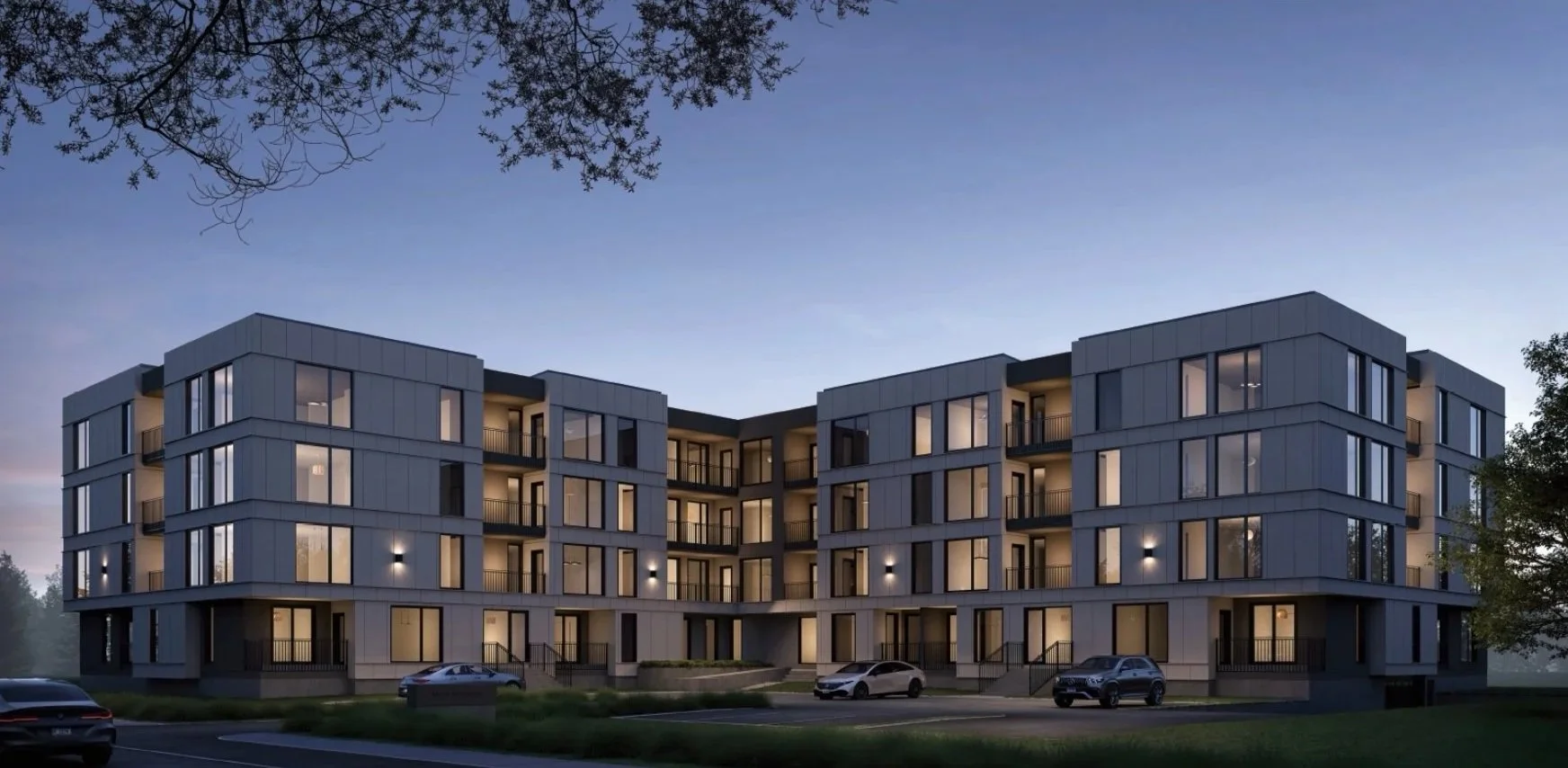 A modern four-story apartment building with a gray exterior, illuminated windows, balconies, and a parking lot with cars in front during twilight.