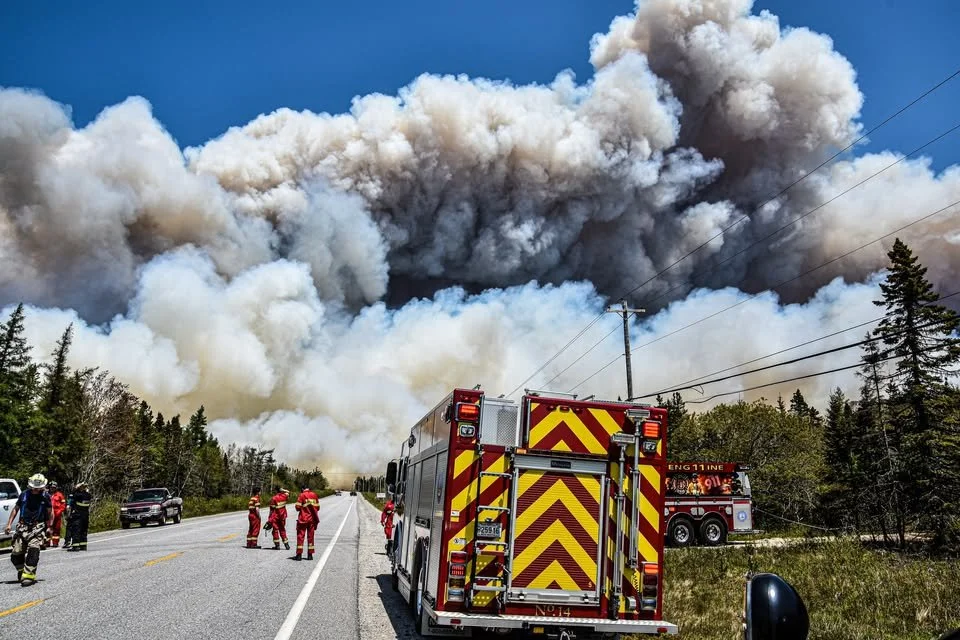 Firefighters and fire trucks responding to a wildfire with large smoke clouds in the sky.