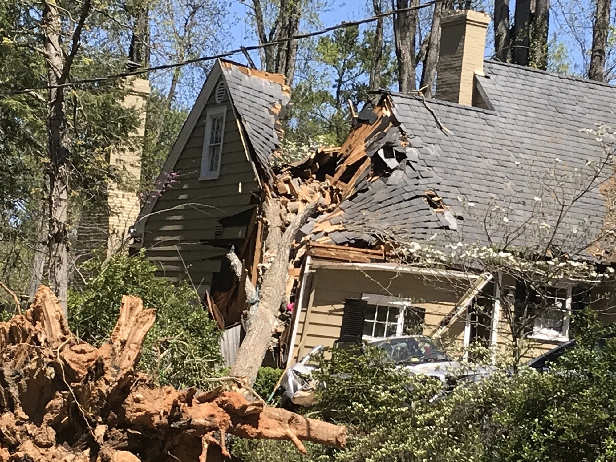 A fallen tree has damaged the roof of a house, with part of the roof torn off and broken wood visible. The house is on a wooded lot with tall trees around.