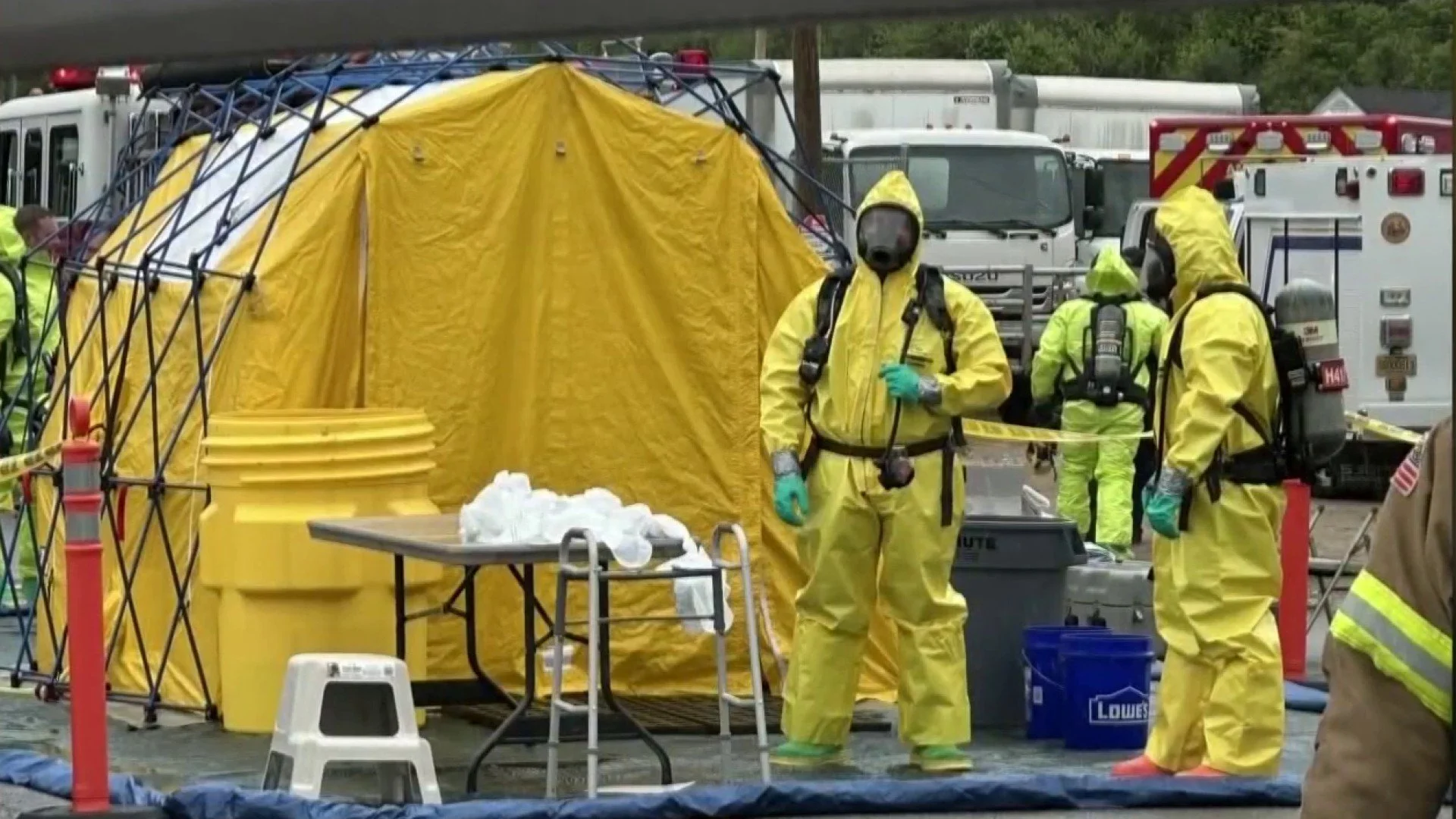 Emergency responders in yellow hazmat suits and gas masks at a scene with a yellow tent and large yellow waste containers, surrounded by trucks and emergency vehicles.