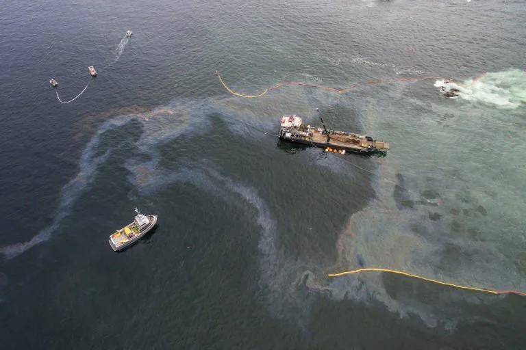Barrel raft with oil containment boom and a vessel nearby on water, possibly involved in oil spill response.